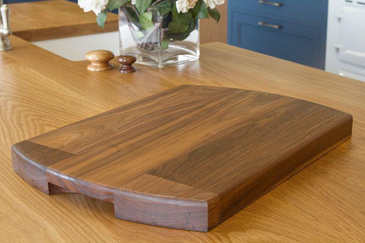 Wooden cutting board with a handle cutout, placed on a wooden countertop. In the background are a vase of flowers and wooden salt and pepper shakers. The setting appears to be a kitchen with blue cabinetry.