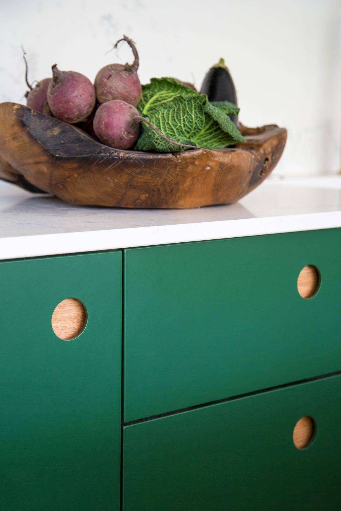 A wooden bowl with beets, cabbage, and an eggplant sits on a white countertop above green cabinets with circular wooden handles.