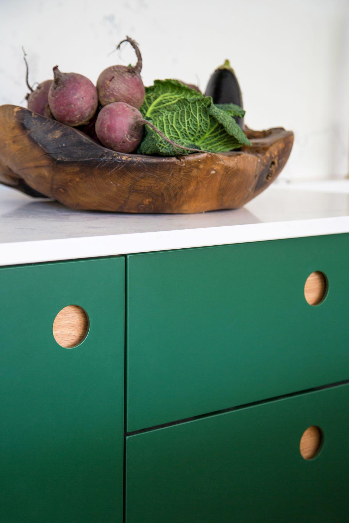 A wooden bowl with beets, cabbage, and an eggplant sits on a white countertop above green cabinets with circular wooden handles.