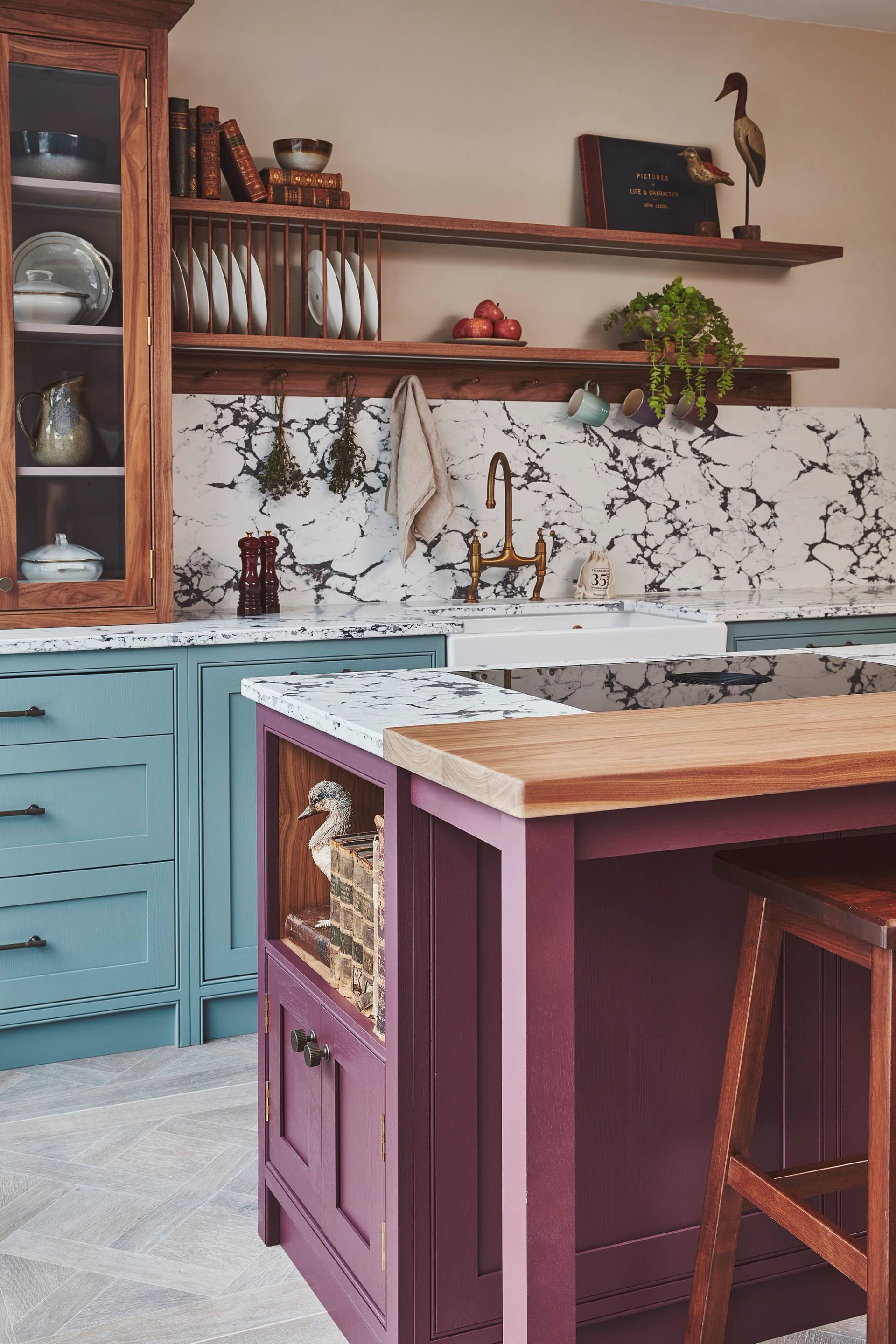 A kitchen featuring a purple island with a wooden countertop, light blue cabinets, and a white marble backsplash. Open shelving displays dishes and a plant. A copper faucet sits above a white farmhouse sink. Decorative items add charm.