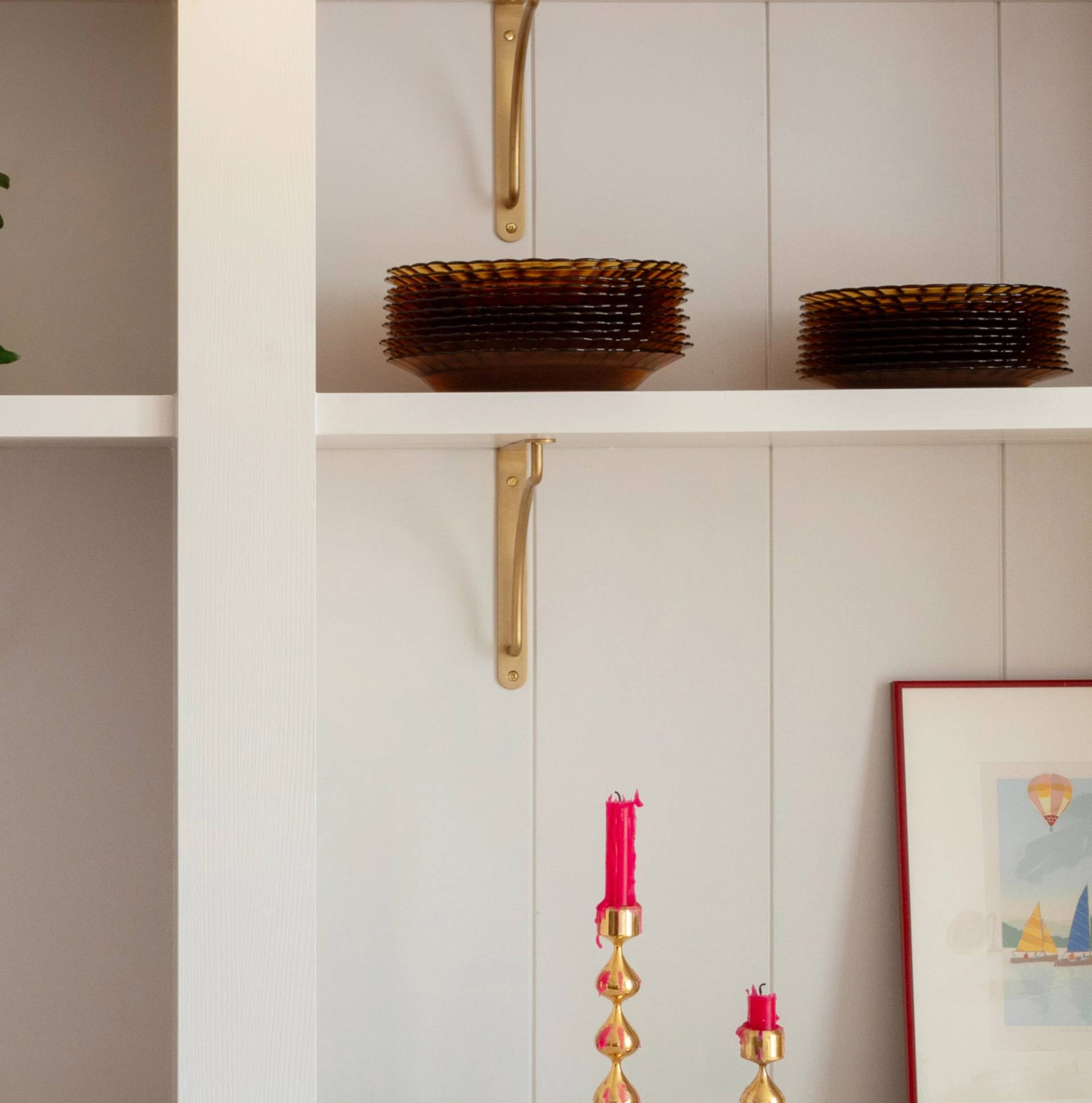 A shelf with stacks of brown glass plates, gold brackets, and part of a framed artwork. Below, two twisted gold candle holders with red candles are visible.