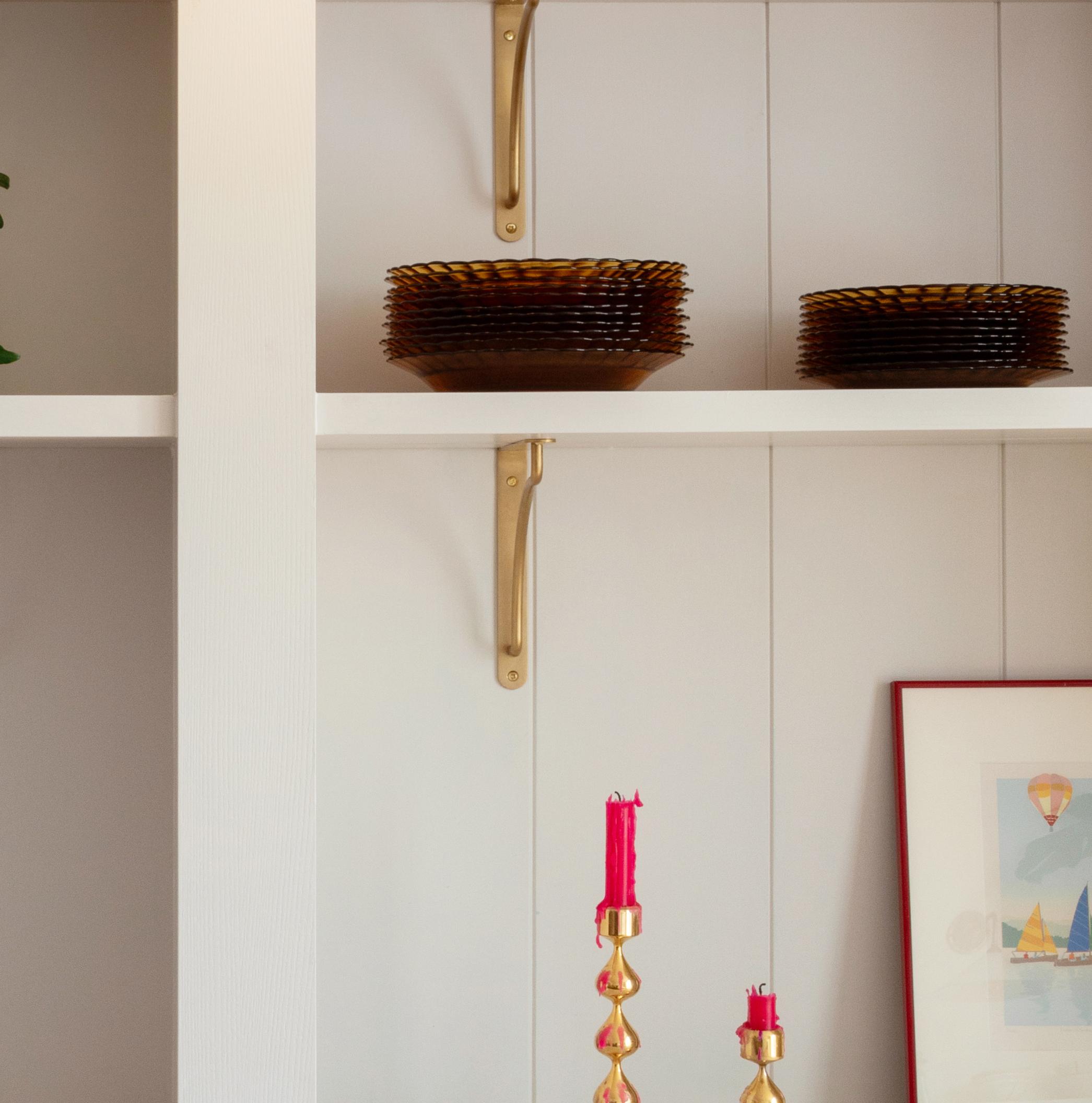 A shelf with stacks of brown glass plates, gold brackets, and part of a framed artwork. Below, two twisted gold candle holders with red candles are visible.