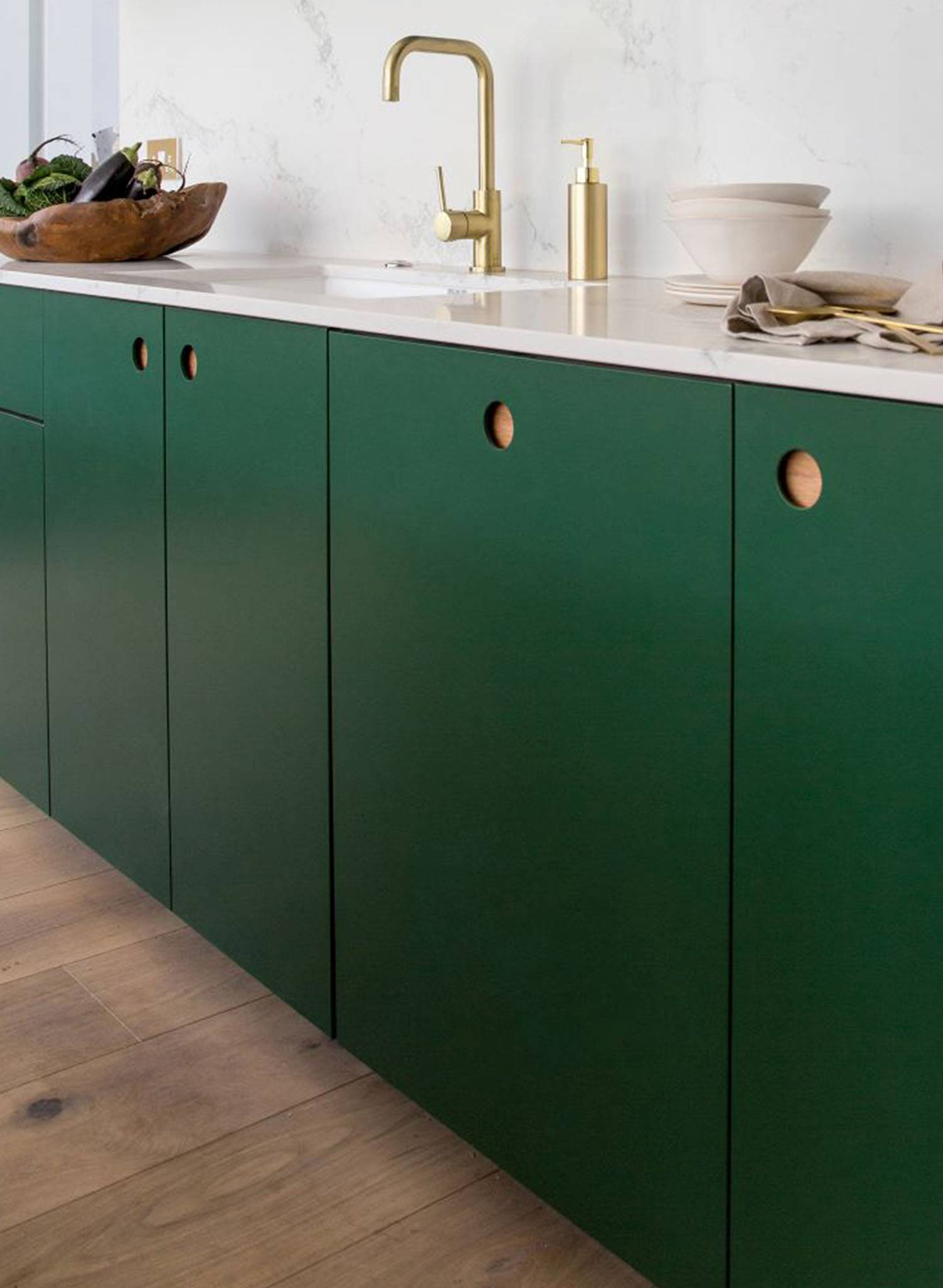 Green kitchen cabinets with round cutout handles, a white countertop, and a gold faucet. A bowl with vegetables and stacked dishes are on the counter, and the floor is light wood.