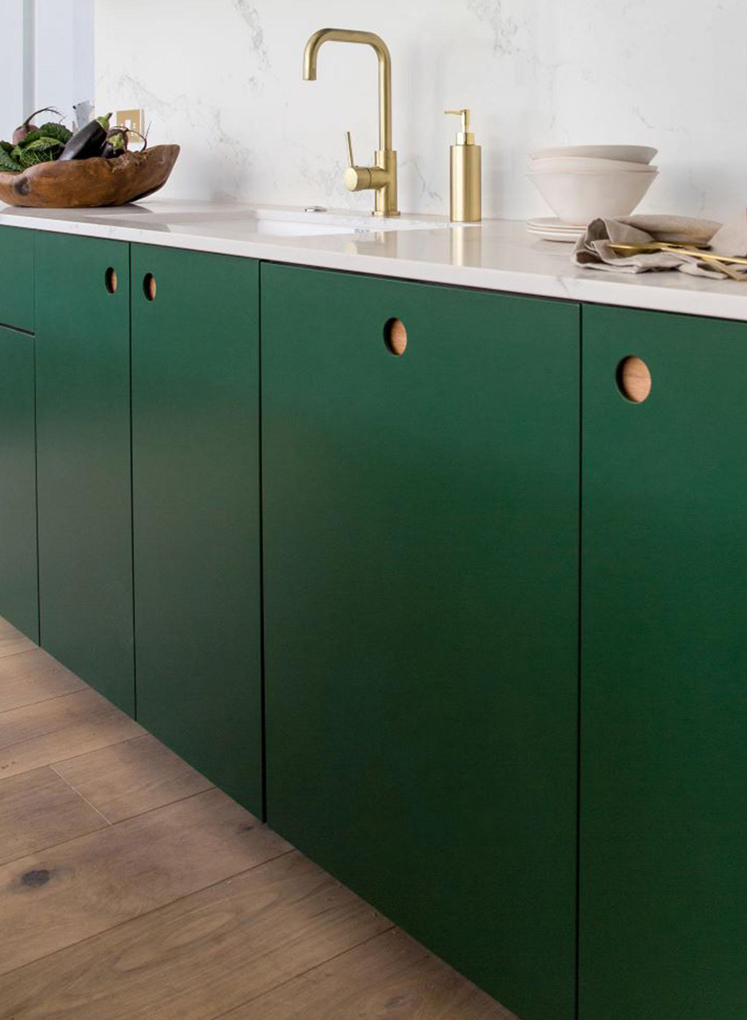 Green kitchen cabinets with round cutout handles, a white countertop, and a gold faucet. A bowl with vegetables and stacked dishes are on the counter, and the floor is light wood.