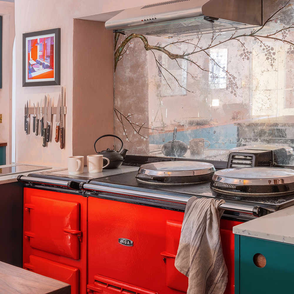 A cozy kitchen scene with a bright red AGA stove, a striped dish towel hanging from the handle, a kettle and mugs on top, knives on a magnetic strip, and a mirror reflecting tree branches and light.