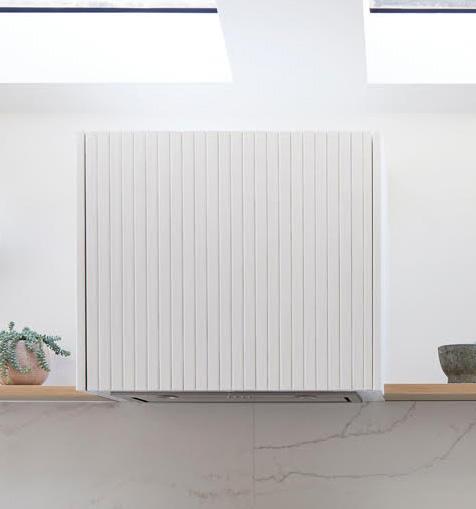 A modern white kitchen hood with vertical grooves is mounted above a light wood shelf. A small potted plant and a bowl sit on the shelf, and skylights above let in natural light.