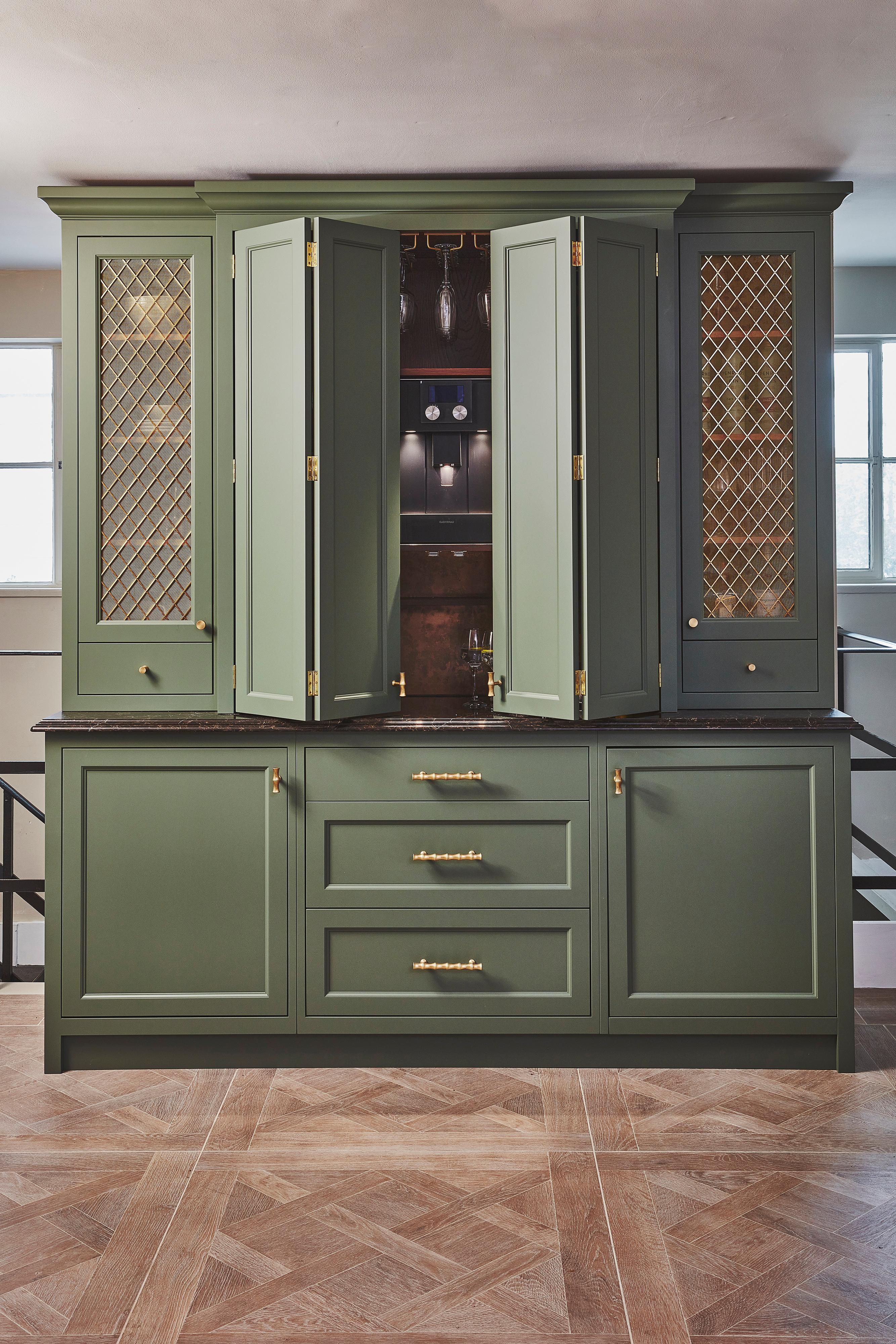 Sage green kitchen cabinets with gold mesh panel inserts, brass handles, and a dark countertop, set against a neutral wall with two windows in the background and geometric patterned wood flooring.