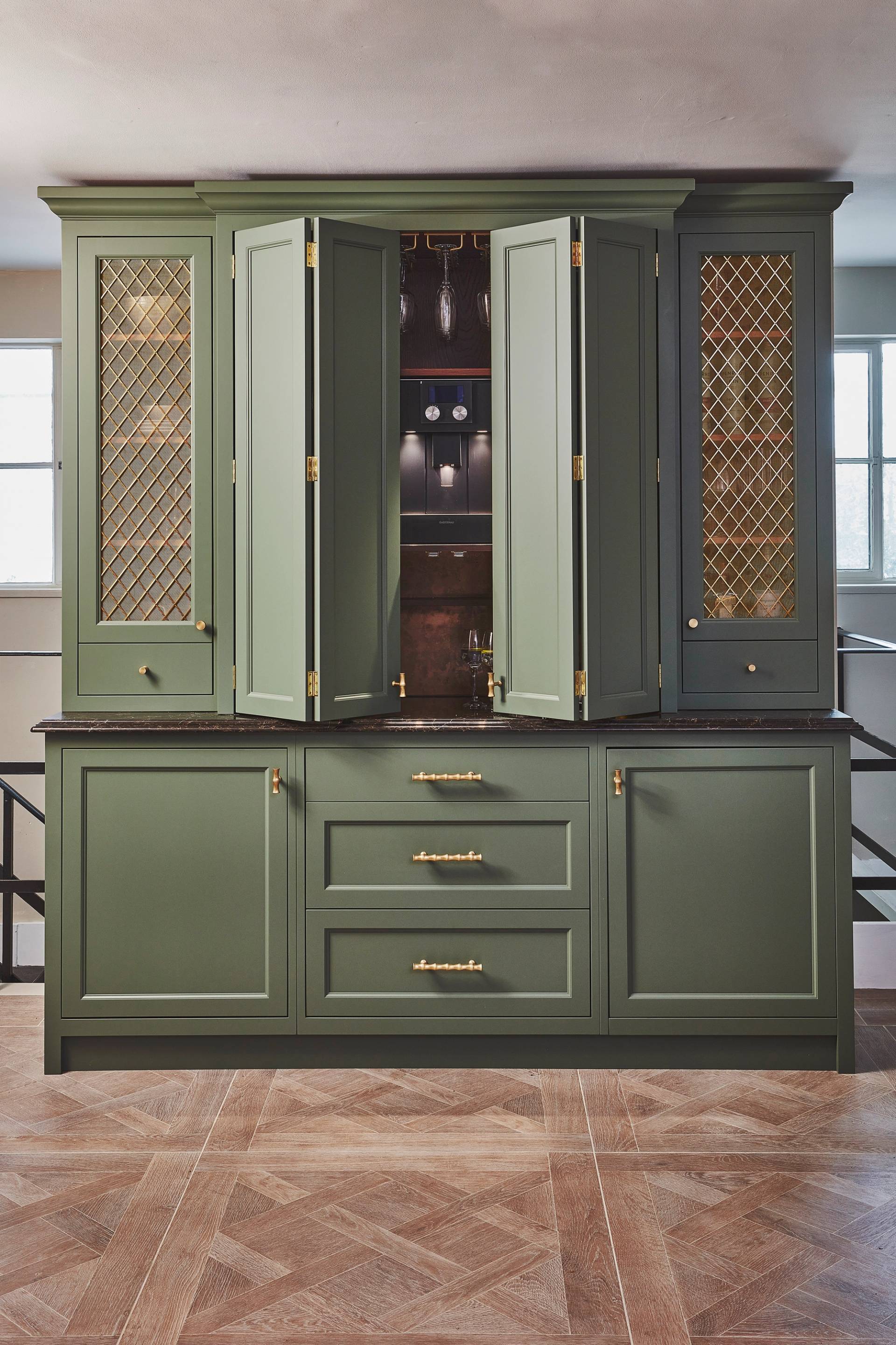 Sage green kitchen cabinets with gold mesh panel inserts, brass handles, and a dark countertop, set against a neutral wall with two windows in the background and geometric patterned wood flooring.