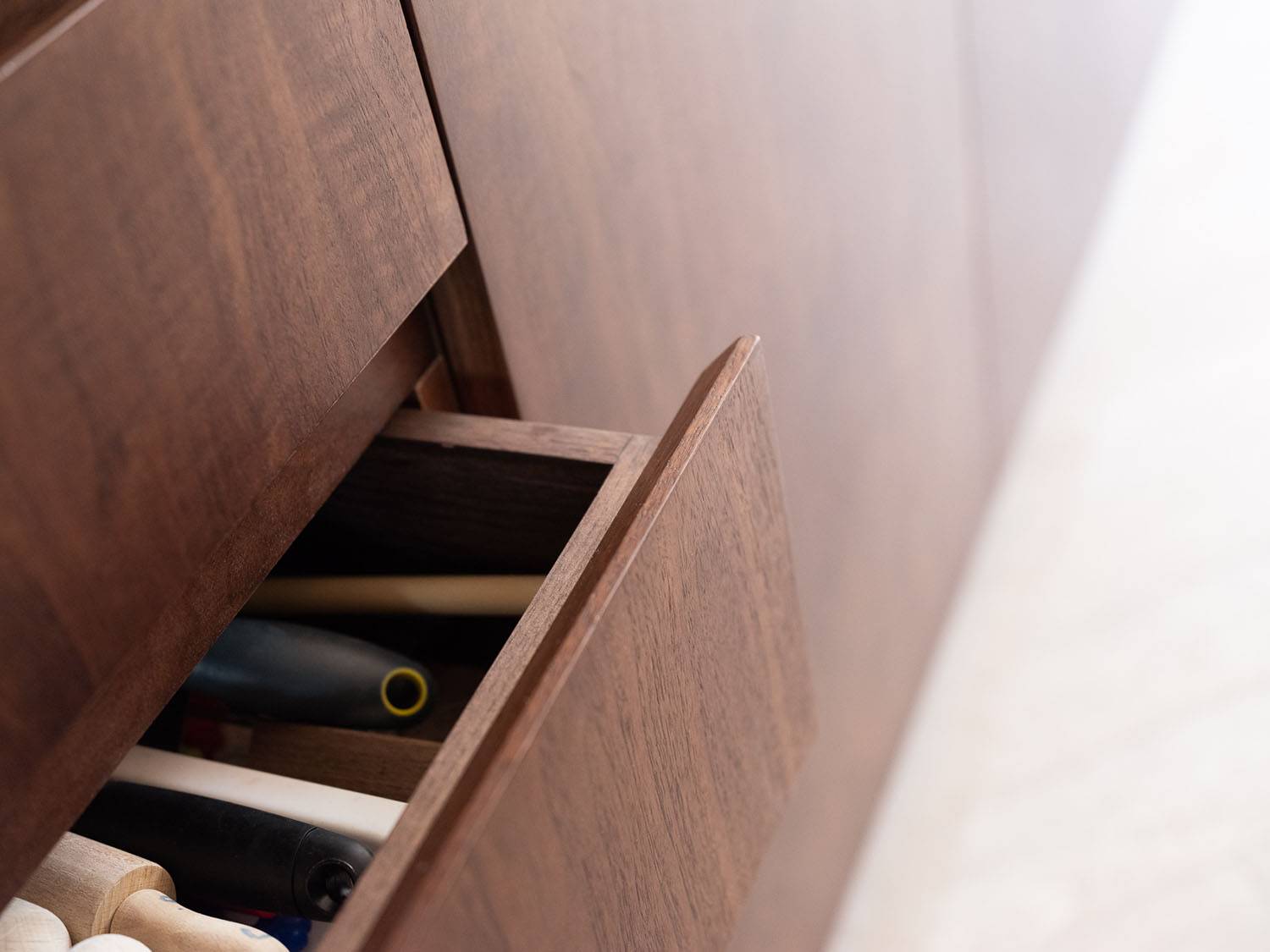 Close-up of a partially open wooden drawer showing assorted tools with handles, such as screwdrivers or similar items, inside a wooden cabinet. The surrounding surfaces are also wood, contributing to a cohesive design.