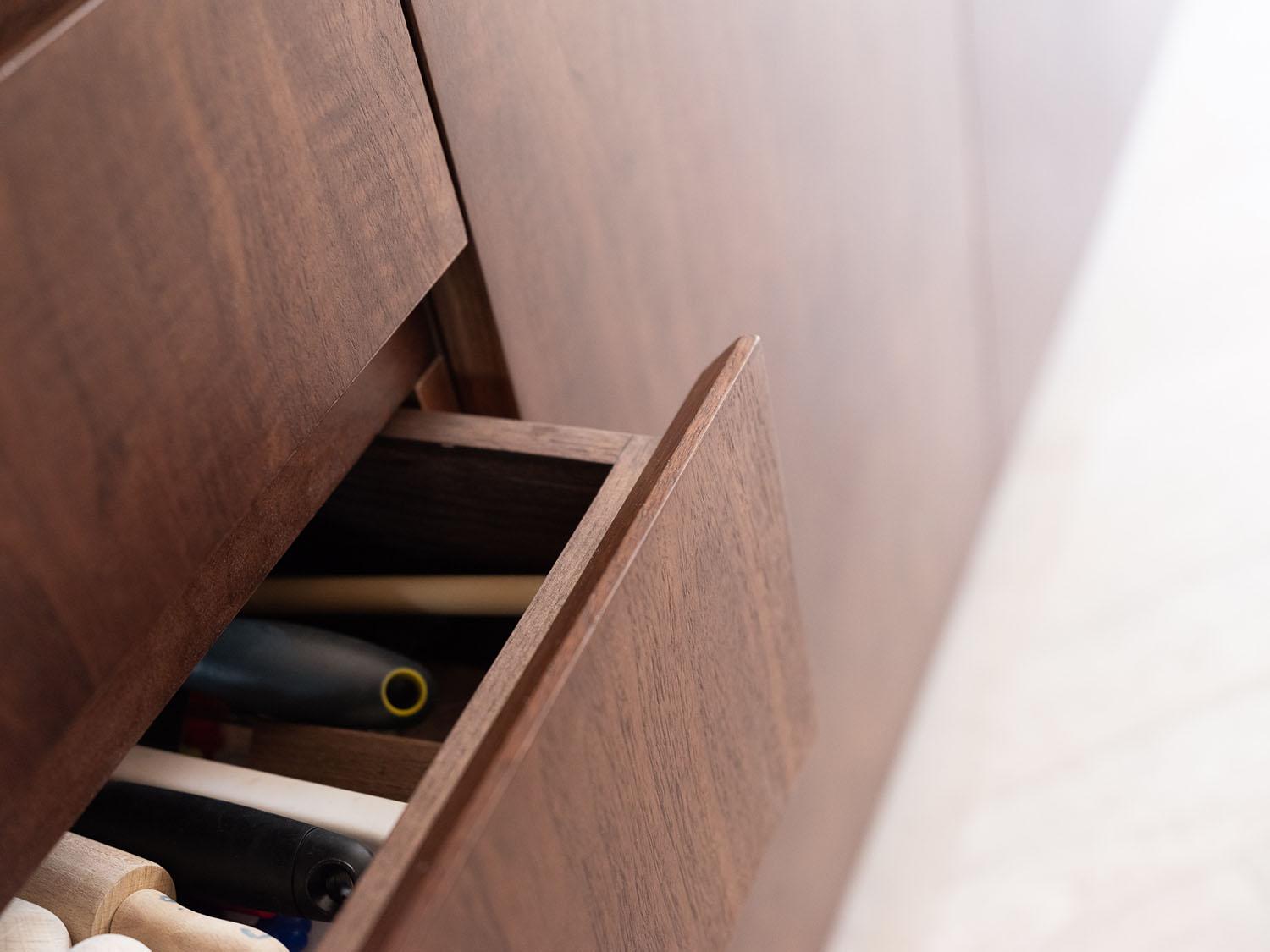 Close-up of a partially open wooden drawer showing assorted tools with handles, such as screwdrivers or similar items, inside a wooden cabinet. The surrounding surfaces are also wood, contributing to a cohesive design.