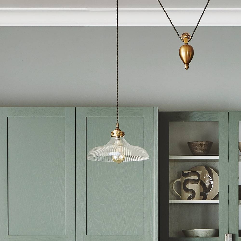 A modern kitchen with green cabinets features a glass pendant light hanging from the ceiling. The cabinets have a mix of solid and glass-fronted doors, displaying decorative pottery. A pulley system is attached to the ceiling.