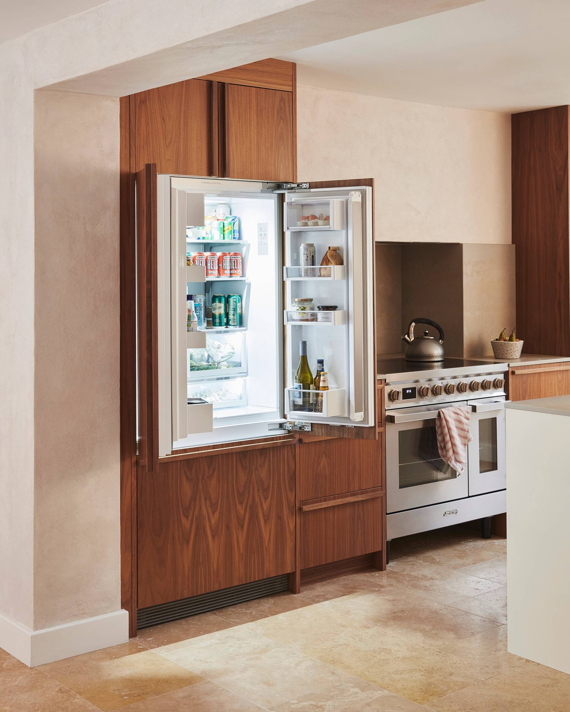 A modern kitchen with wooden cabinets, a stainless steel oven, and a white countertop. A kettle sits on the stovetop, and a small plant is placed on the counter next to the oven. The floor is made of light beige tiles.