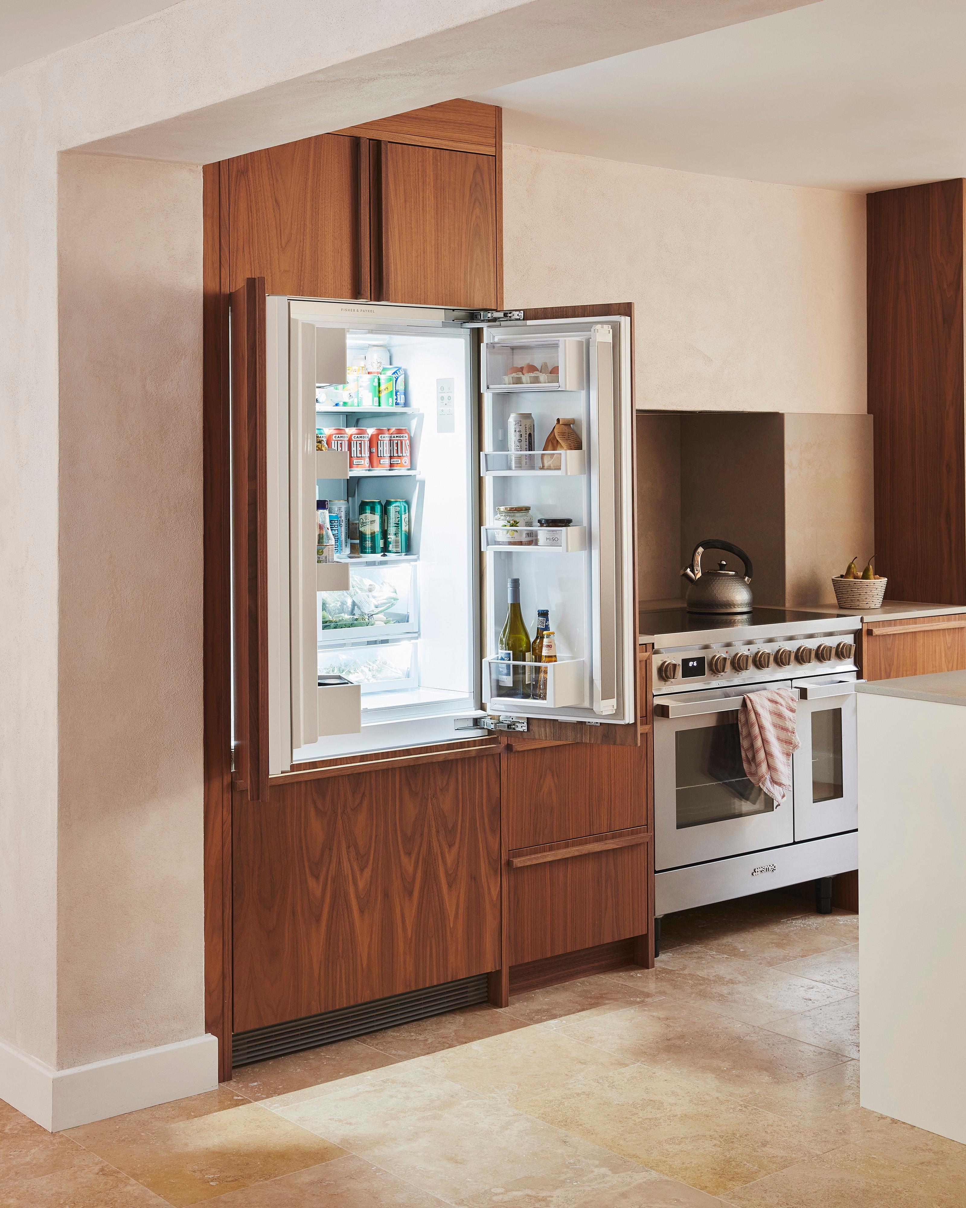 A modern kitchen with wooden cabinets, a stainless steel oven, and a white countertop. A kettle sits on the stovetop, and a small plant is placed on the counter next to the oven. The floor is made of light beige tiles.