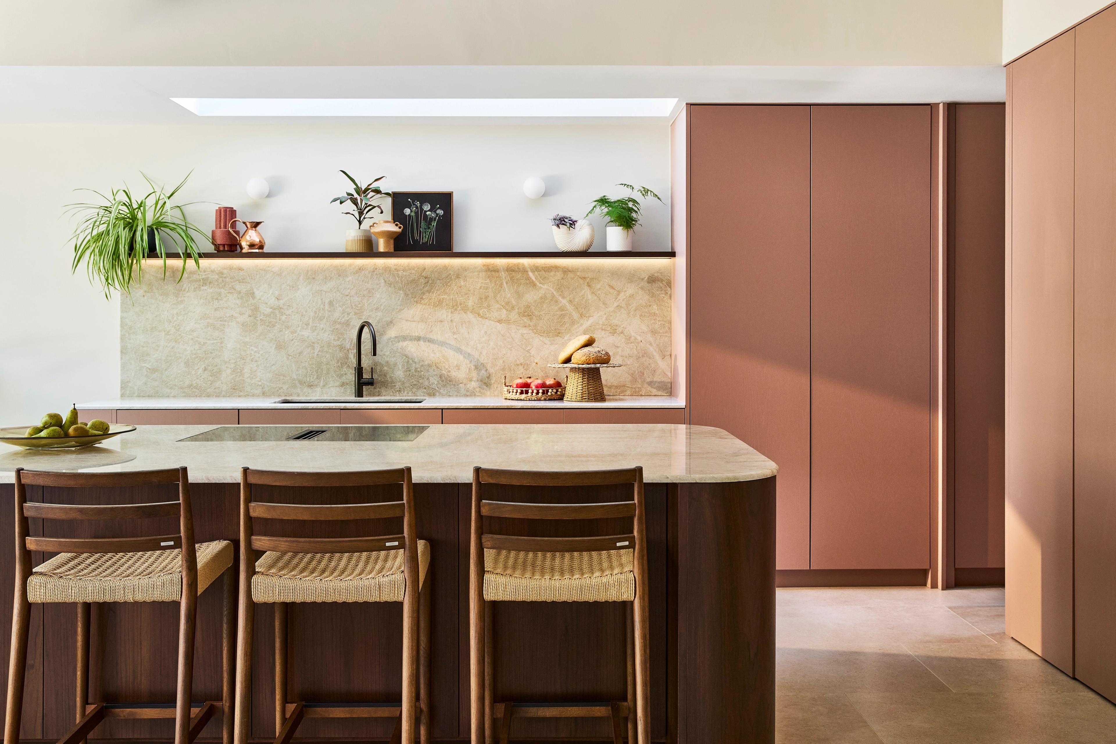 Modern kitchen with wooden cabinets, a white oven, a silver kettle, open shelves with spices and plants, a white island, beige tiled floor, and natural light from a window.