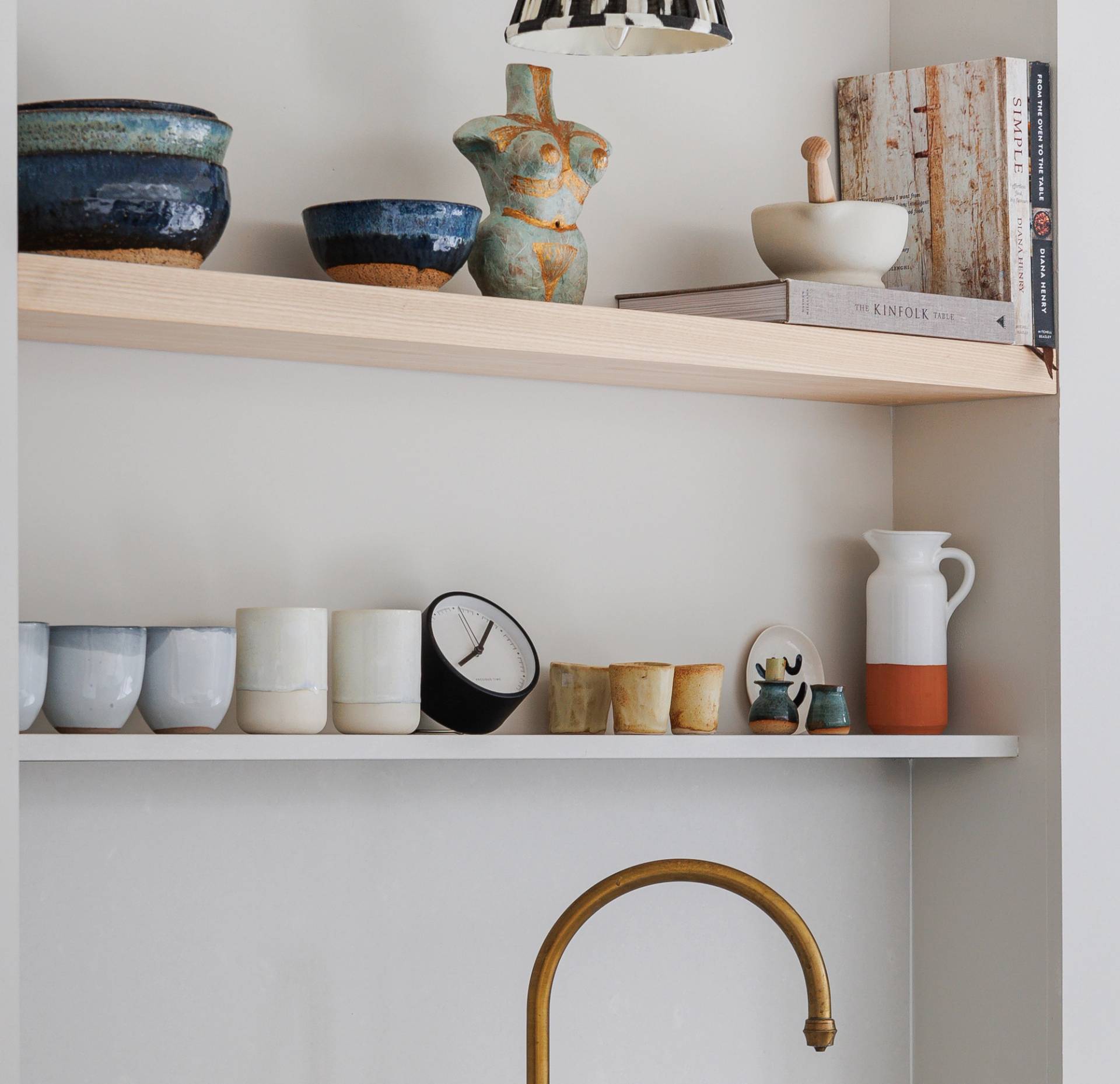 Open shelving displays ceramic bowls, cups, and mugs, along with a clock, decorative figurines, books, a mortar and pestle, and a pitcher above a brass kitchen faucet, against a light wall.