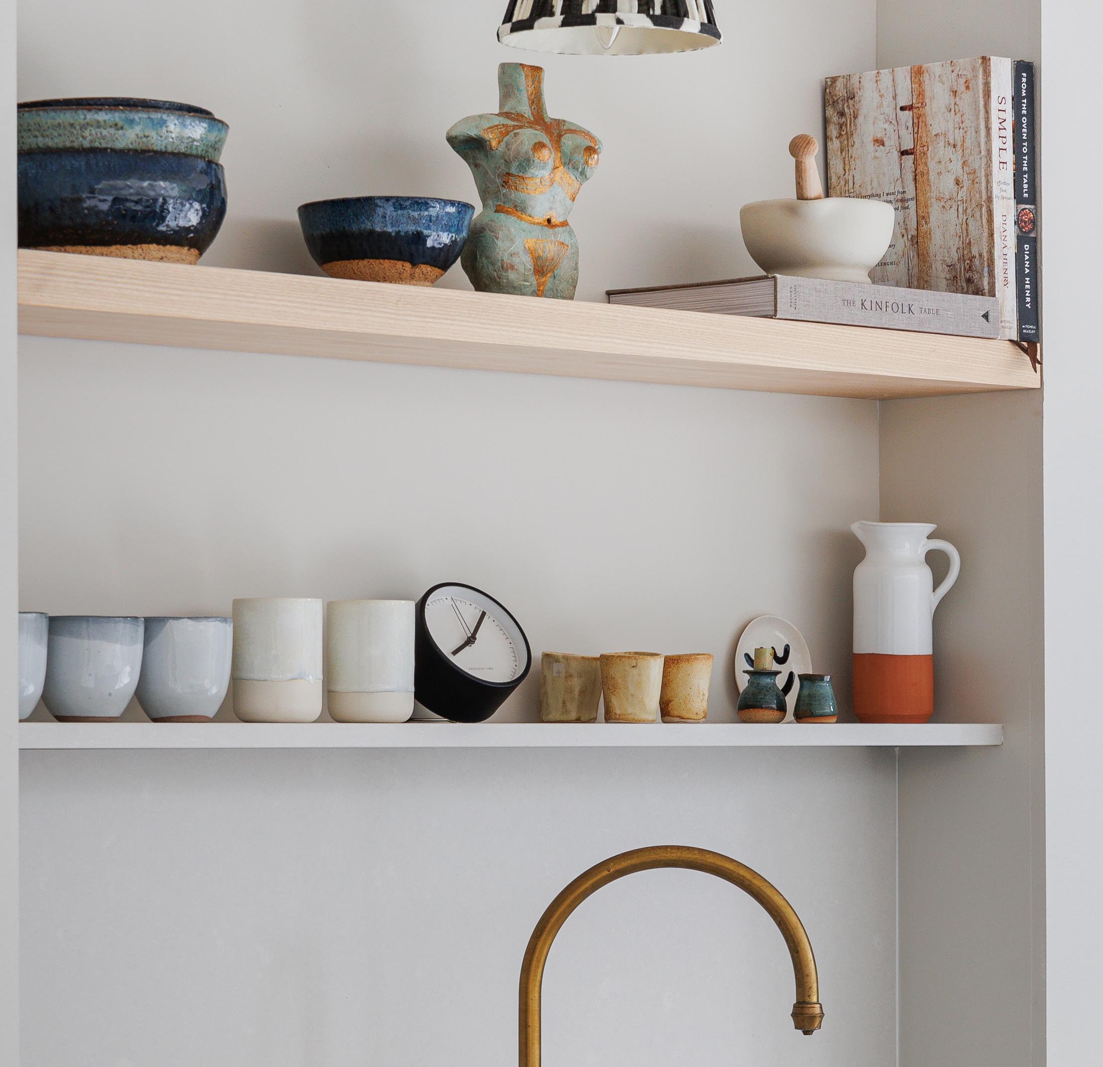 Open shelving displays ceramic bowls, cups, and mugs, along with a clock, decorative figurines, books, a mortar and pestle, and a pitcher above a brass kitchen faucet, against a light wall.