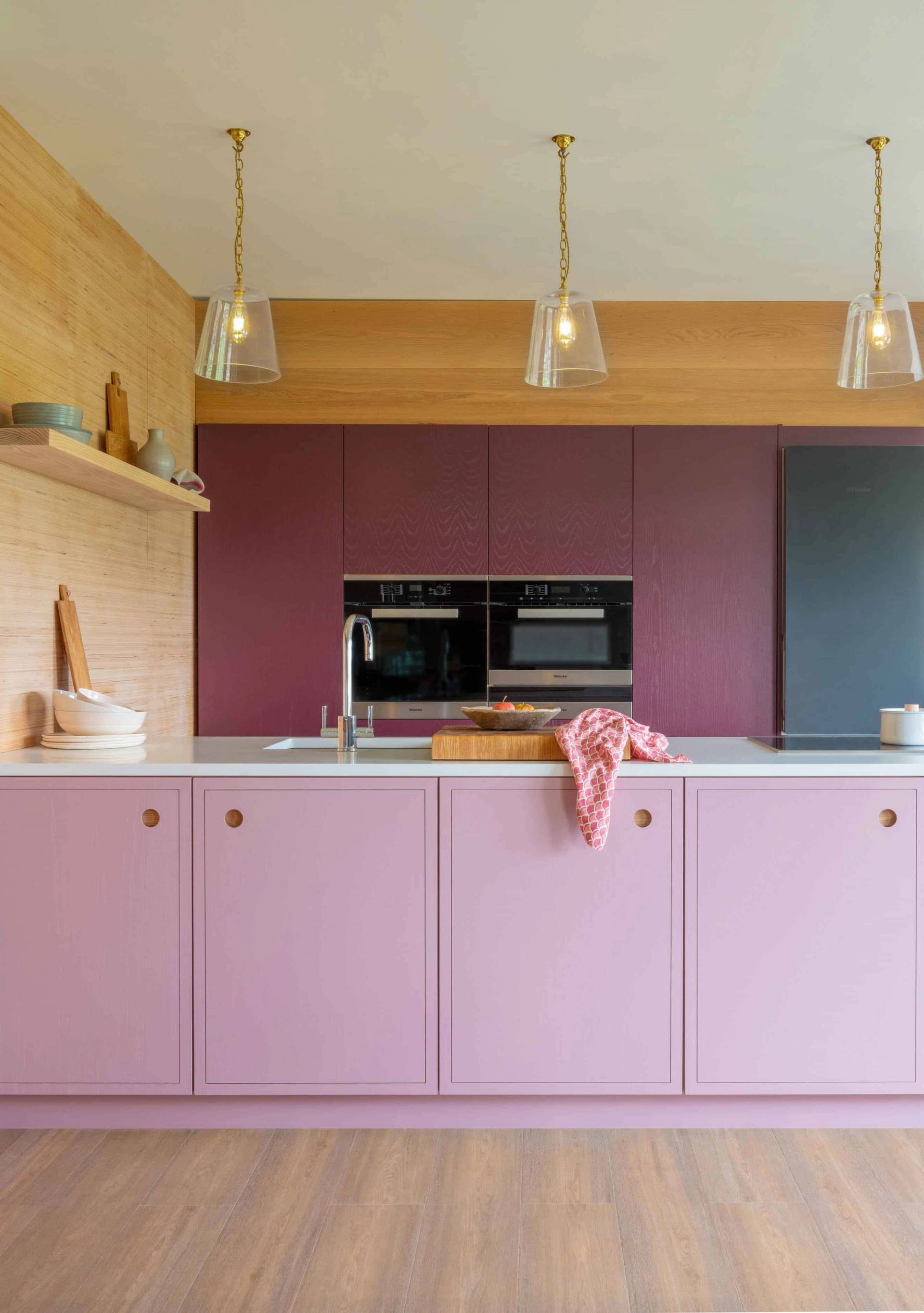A modern kitchen with pink cabinets, a wooden backsplash, and pendant lights. The countertop has a sink and a cutting board. The background features built-in ovens with dark doors. The floor is light wood, complementing the warm, vibrant decor.