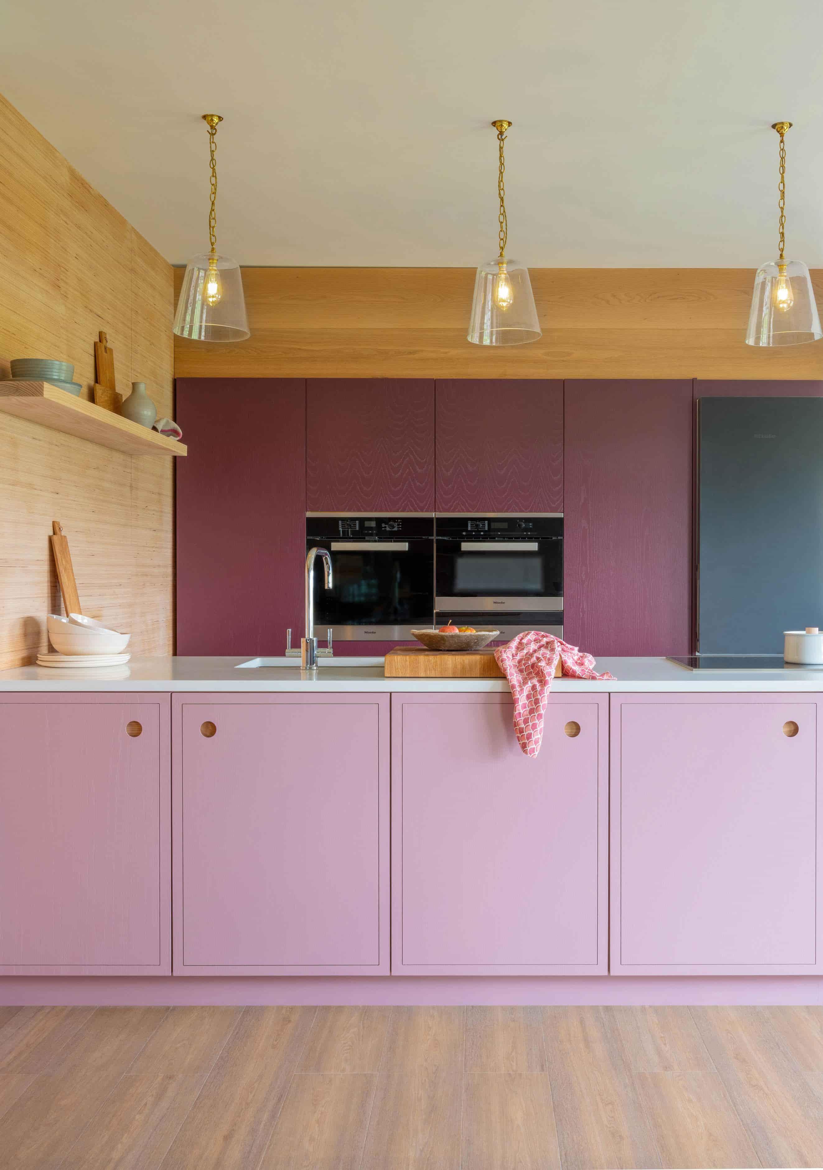 A modern kitchen with pink cabinets, a wooden backsplash, and pendant lights. The countertop has a sink and a cutting board. The background features built-in ovens with dark doors. The floor is light wood, complementing the warm, vibrant decor.