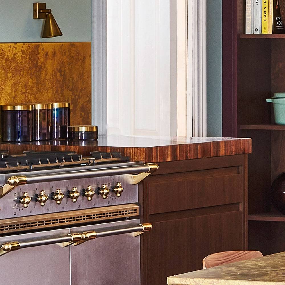 A modern kitchen with a dark wood cabinet, a stainless steel stove with gold accents, colorful containers on the counter, and cookbooks on a shelf in the background.