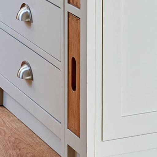 Close-up of a light gray cabinet with metal cup drawer handles. A wooden breadboard with a hand slot is stored vertically in a narrow slot beside the drawers. The cabinet stands on a wood floor.