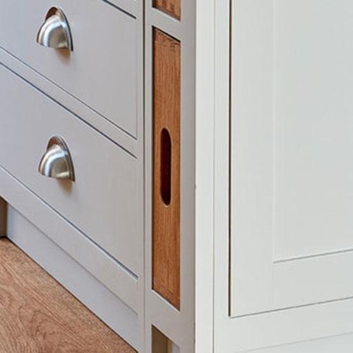 Close-up of a light gray cabinet with metal cup drawer handles. A wooden breadboard with a hand slot is stored vertically in a narrow slot beside the drawers. The cabinet stands on a wood floor.
