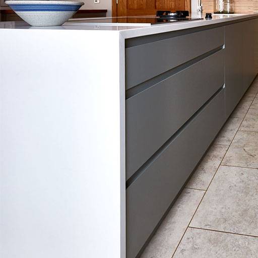 A modern kitchen with sleek, handleless gray cabinets and white countertops. A ceramic bowl with a blue rim sits on the counter, and the floor is tiled in a light stone pattern.