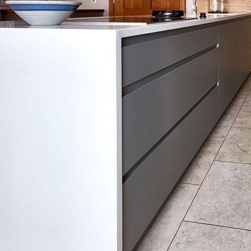 A modern kitchen with sleek, handleless gray cabinets and white countertops. A ceramic bowl with a blue rim sits on the counter, and the floor is tiled in a light stone pattern.