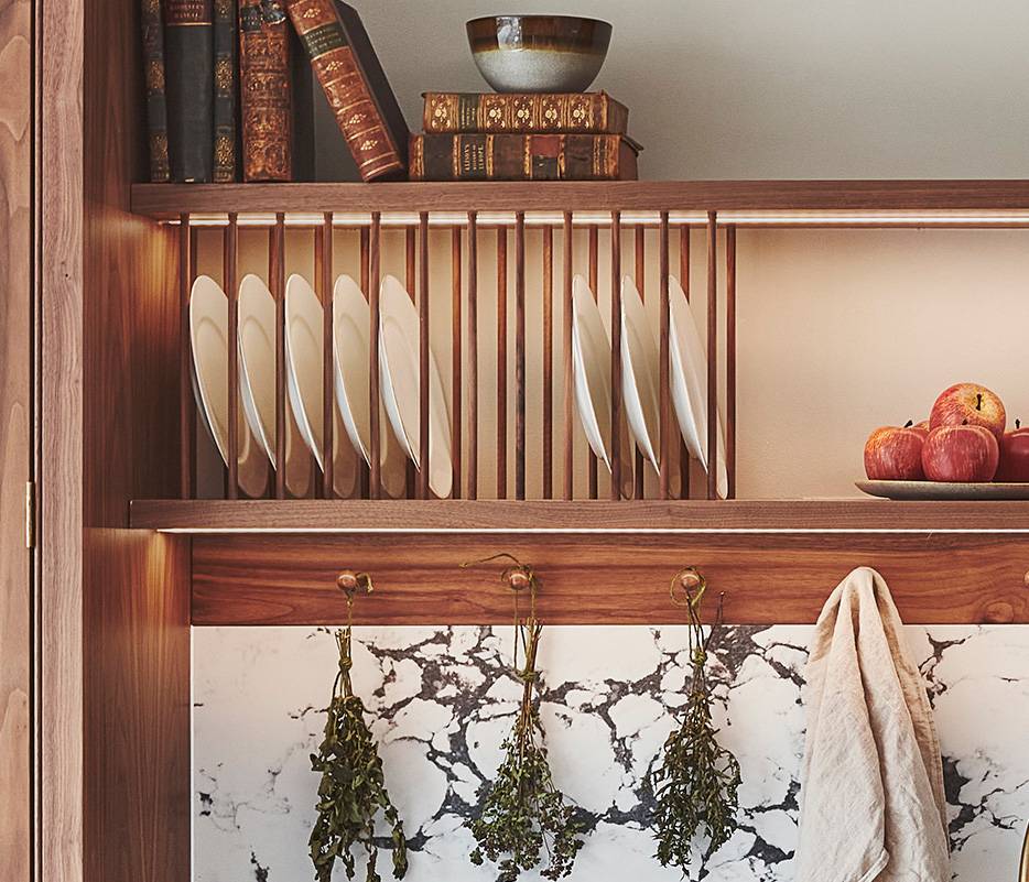 Wooden kitchen shelves with plates, bowls, and vintage books. A bowl of apples rests on the middle shelf. Below, sprigs of dried herbs hang above a marble countertop with a dish towel on the side