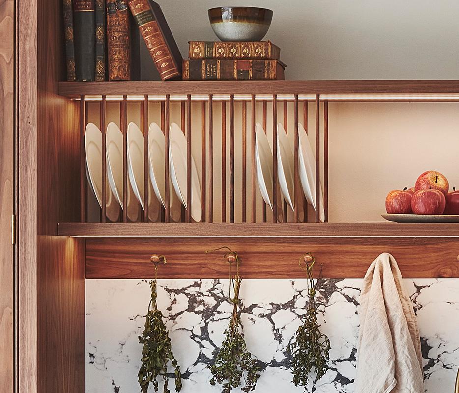 Wooden kitchen shelves with plates, bowls, and vintage books. A bowl of apples rests on the middle shelf. Below, sprigs of dried herbs hang above a marble countertop with a dish towel on the side