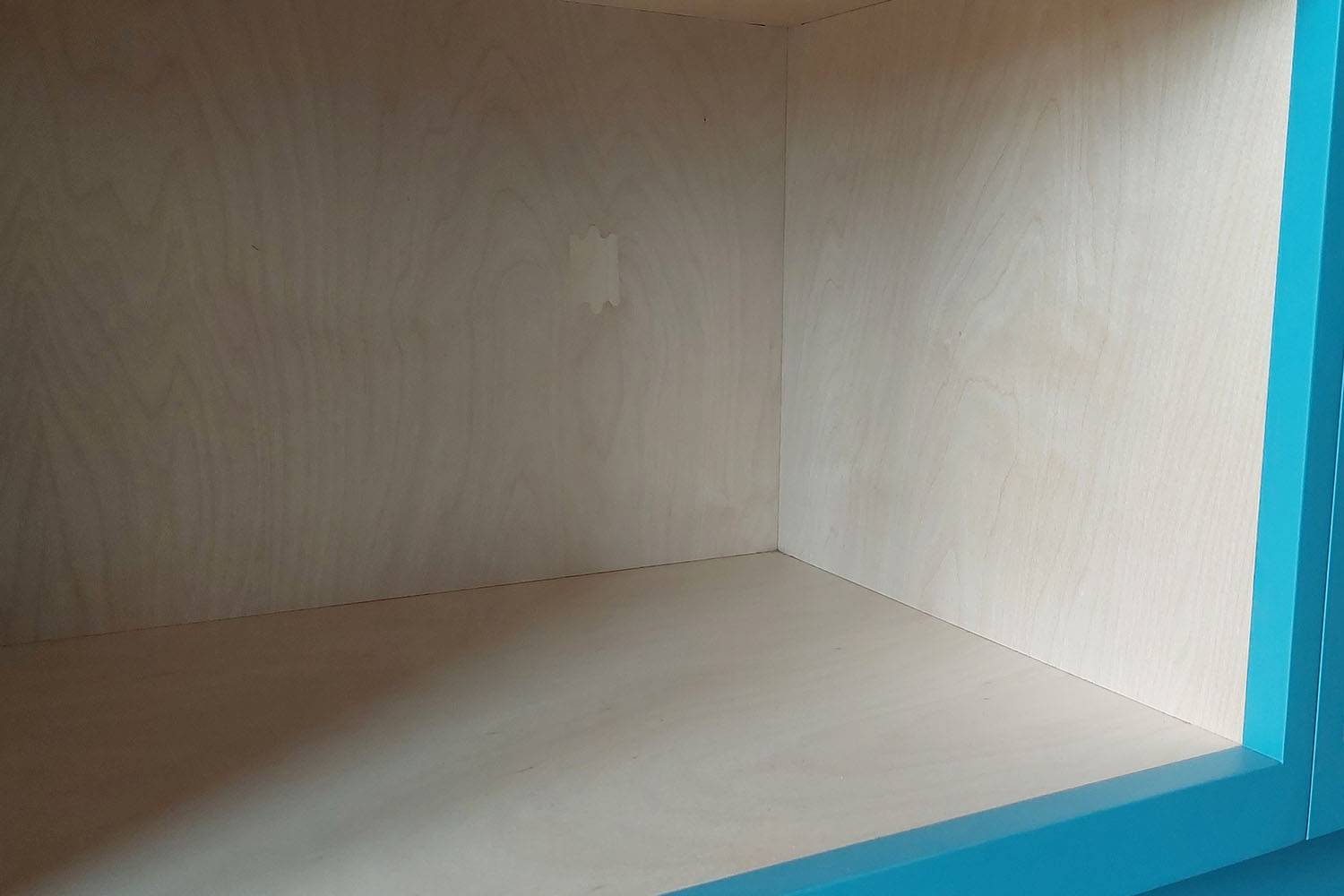 An empty wooden shelf with a light wood finish and a blue painted frame. The interior of the shelf shows a smooth surface with a small patch on the back wall. The shelf is brightly lit, highlighting the wood grain.