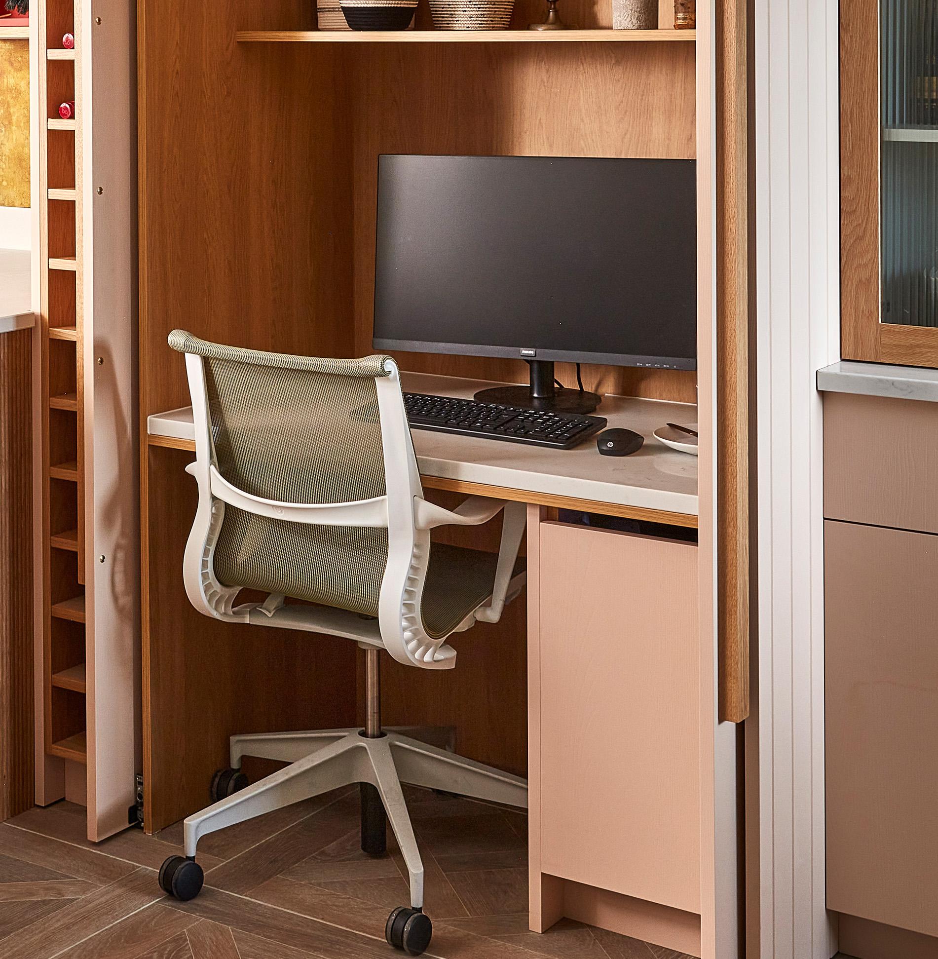 Home office nook with a tan desk and chair. A computer monitor and mouse sit on the desk. Wooden shelves, some with decorative items, are above. Theres a vertical shelf to the left, and wood-patterned flooring below.