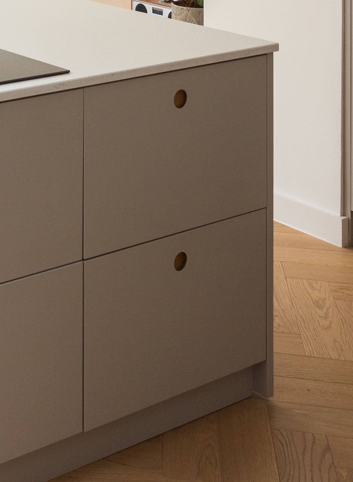 A modern kitchen cabinet with matte beige drawers and round wooden handles, set against a light wood floor and white wall. Part of a black stovetop is visible on the countertop.