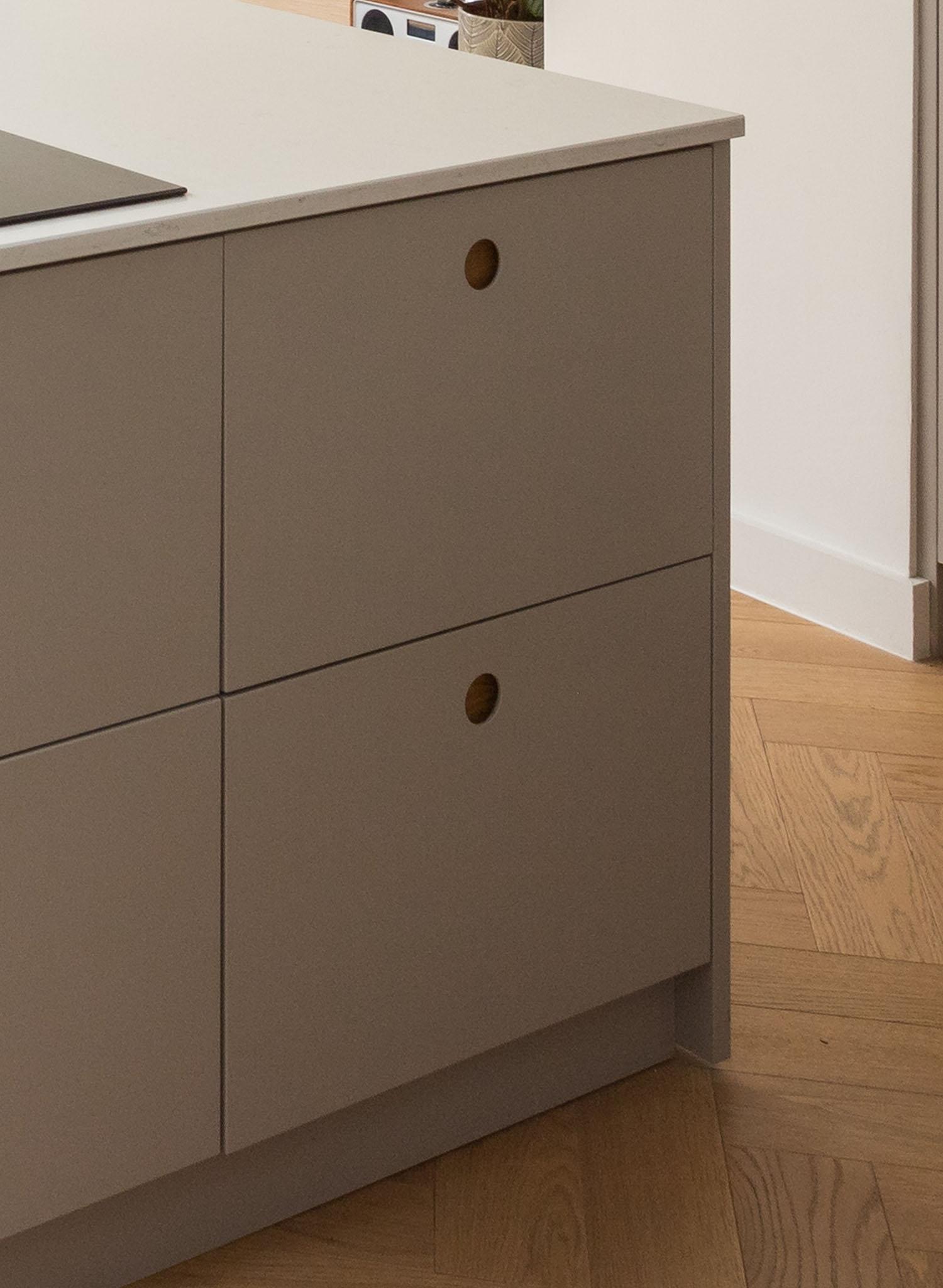 A modern kitchen cabinet with matte beige drawers and round wooden handles, set against a light wood floor and white wall. Part of a black stovetop is visible on the countertop.