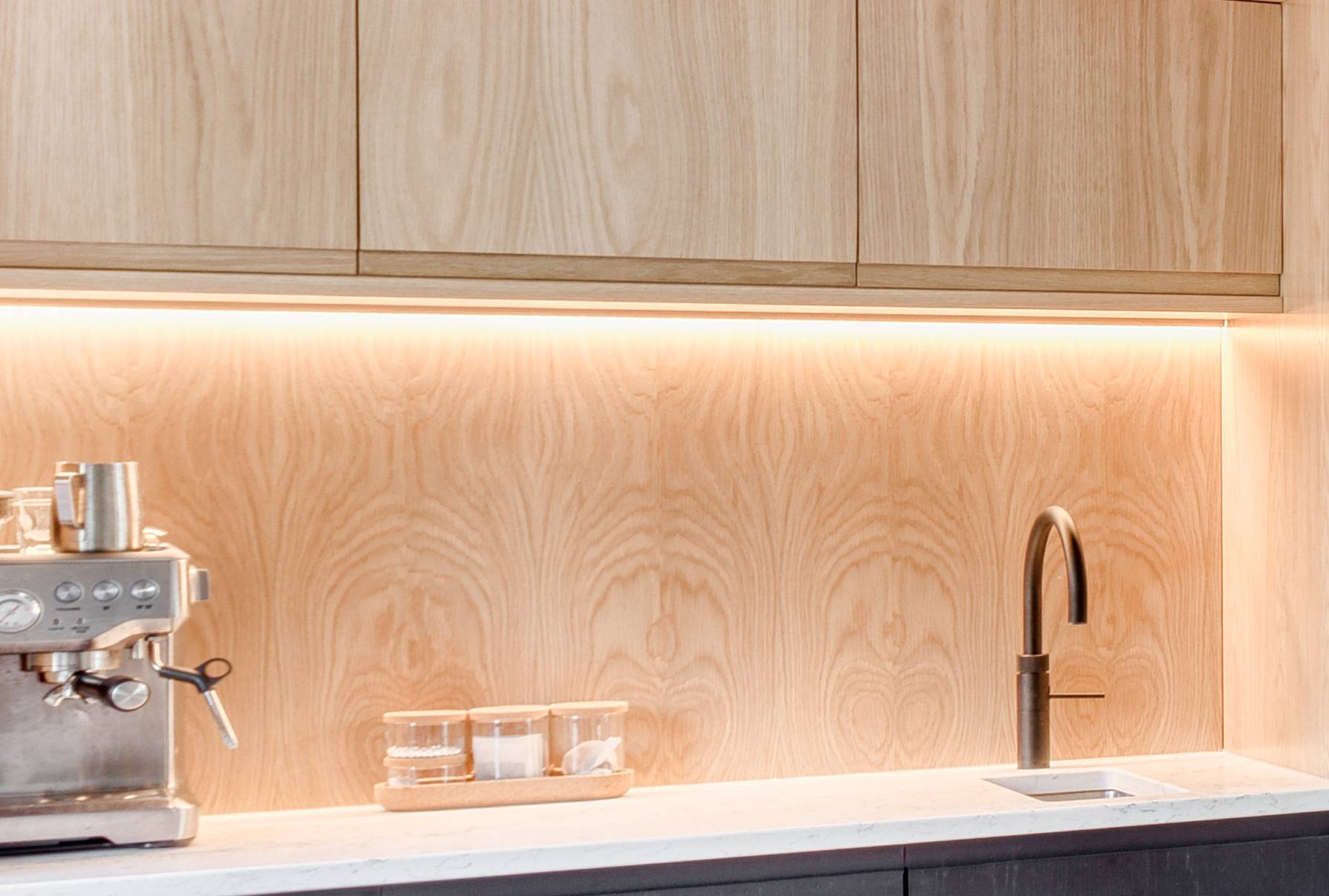 A modern kitchen counter with a silver espresso machine, glass containers, and a sleek faucet beneath light wood cabinets and a matching wood backsplash with under-cabinet lighting.