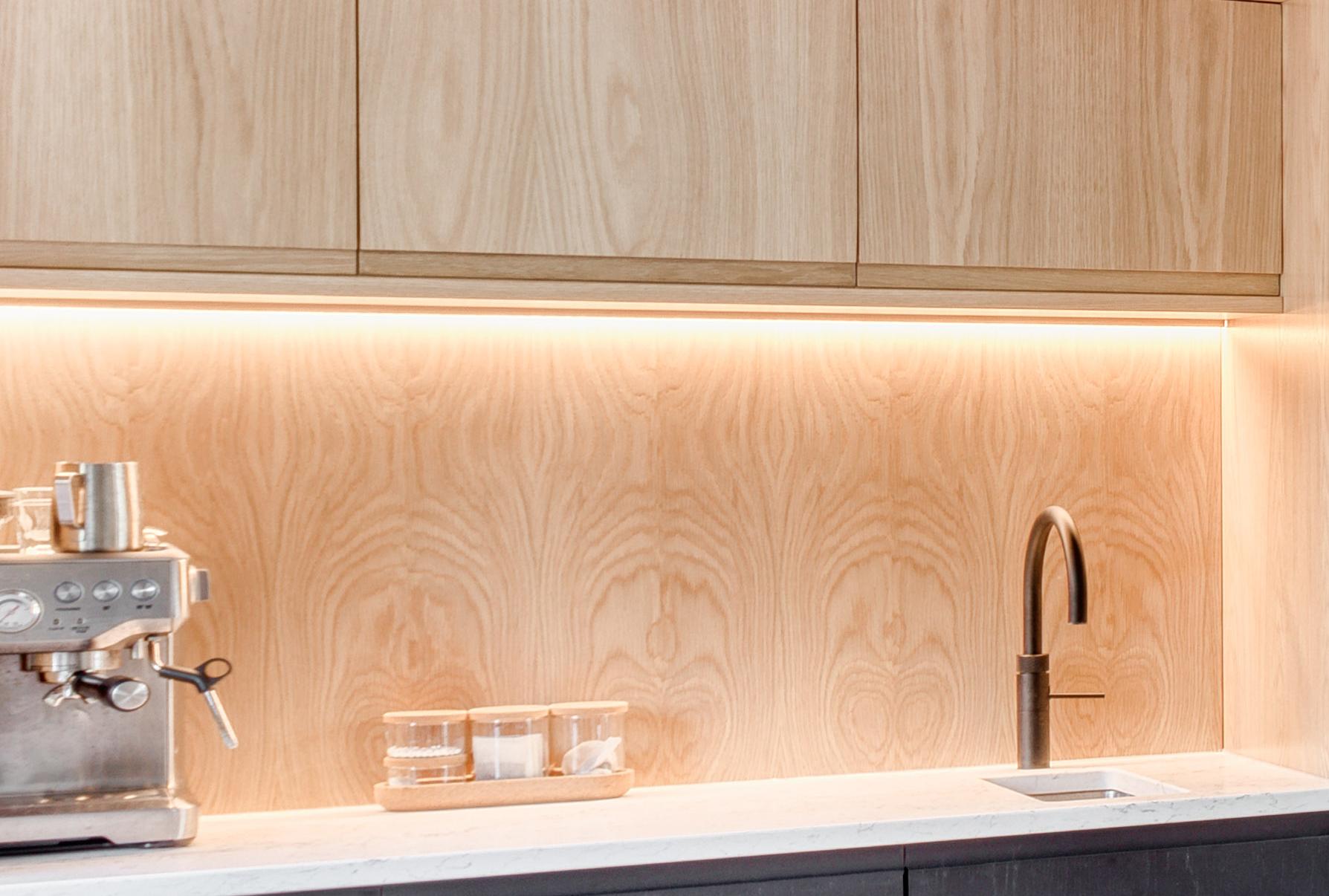 A modern kitchen counter with a silver espresso machine, glass containers, and a sleek faucet beneath light wood cabinets and a matching wood backsplash with under-cabinet lighting.