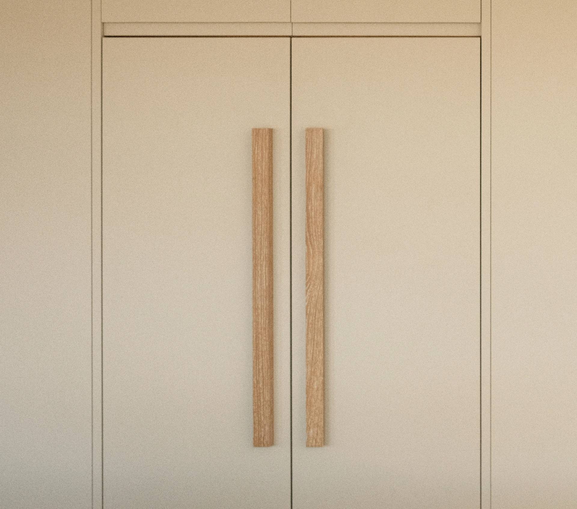 A pair of minimal, beige cabinet doors with long, vertical wooden handles in the center, blending seamlessly into a neutral-colored wall.