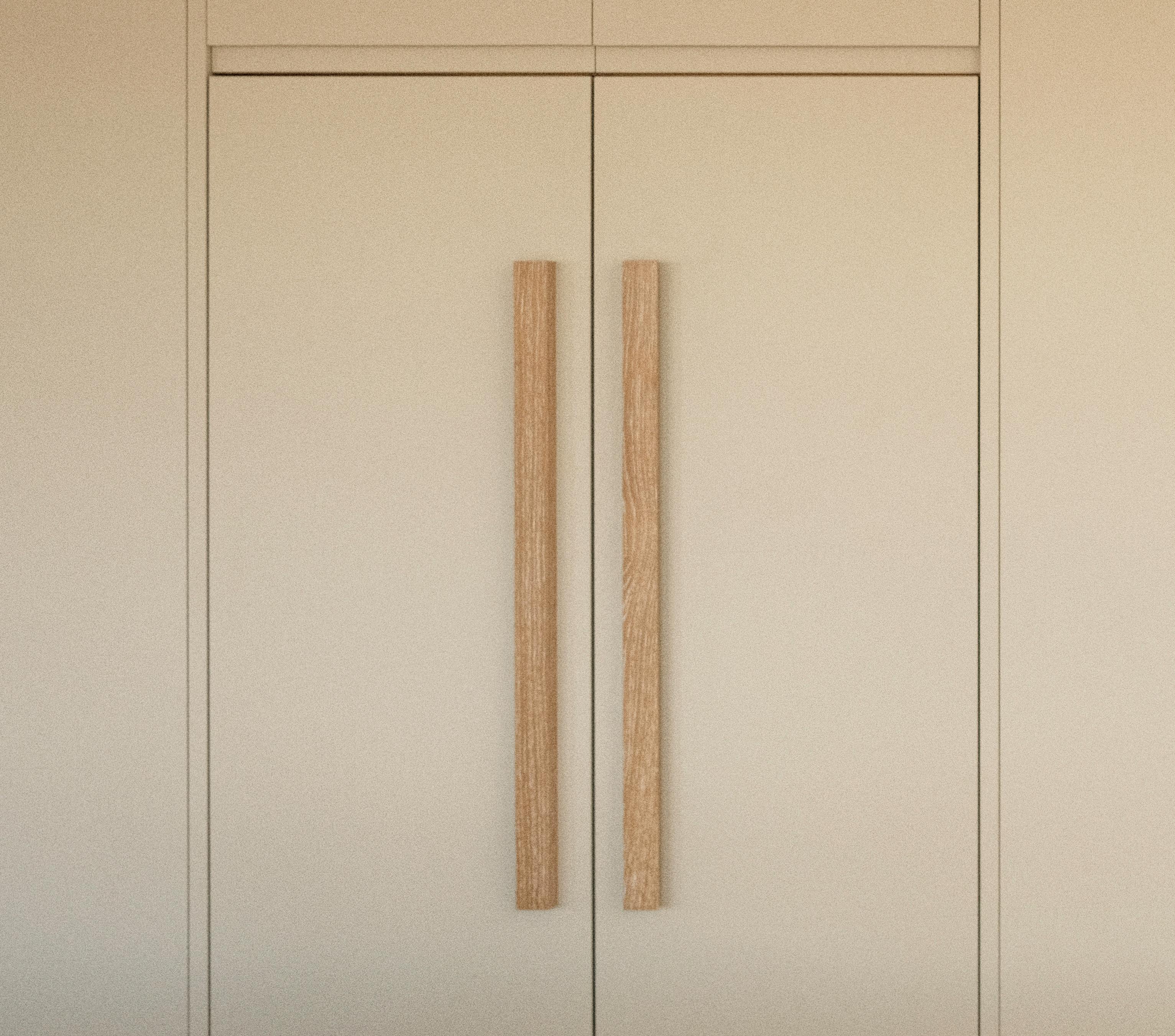 A pair of minimal, beige cabinet doors with long, vertical wooden handles in the center, blending seamlessly into a neutral-colored wall.