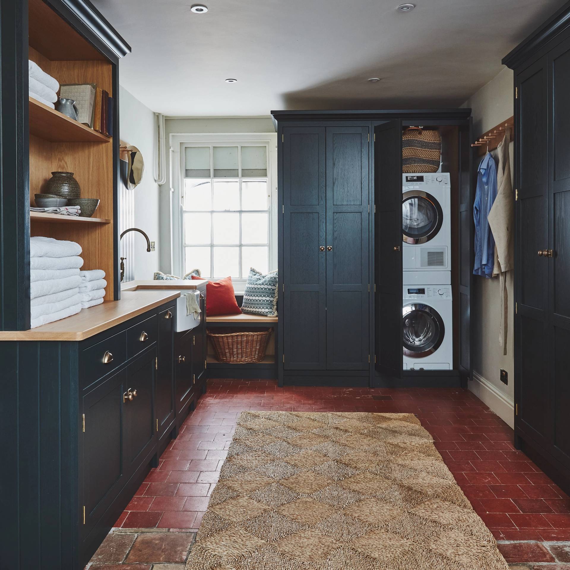 Laundry room with dark cabinets, a stacked washer and dryer, brick floor, and a woven rug. Wooden countertop holds folded towels. A window seat with cushions is in the corner, and there are hangers with shirts on the right.