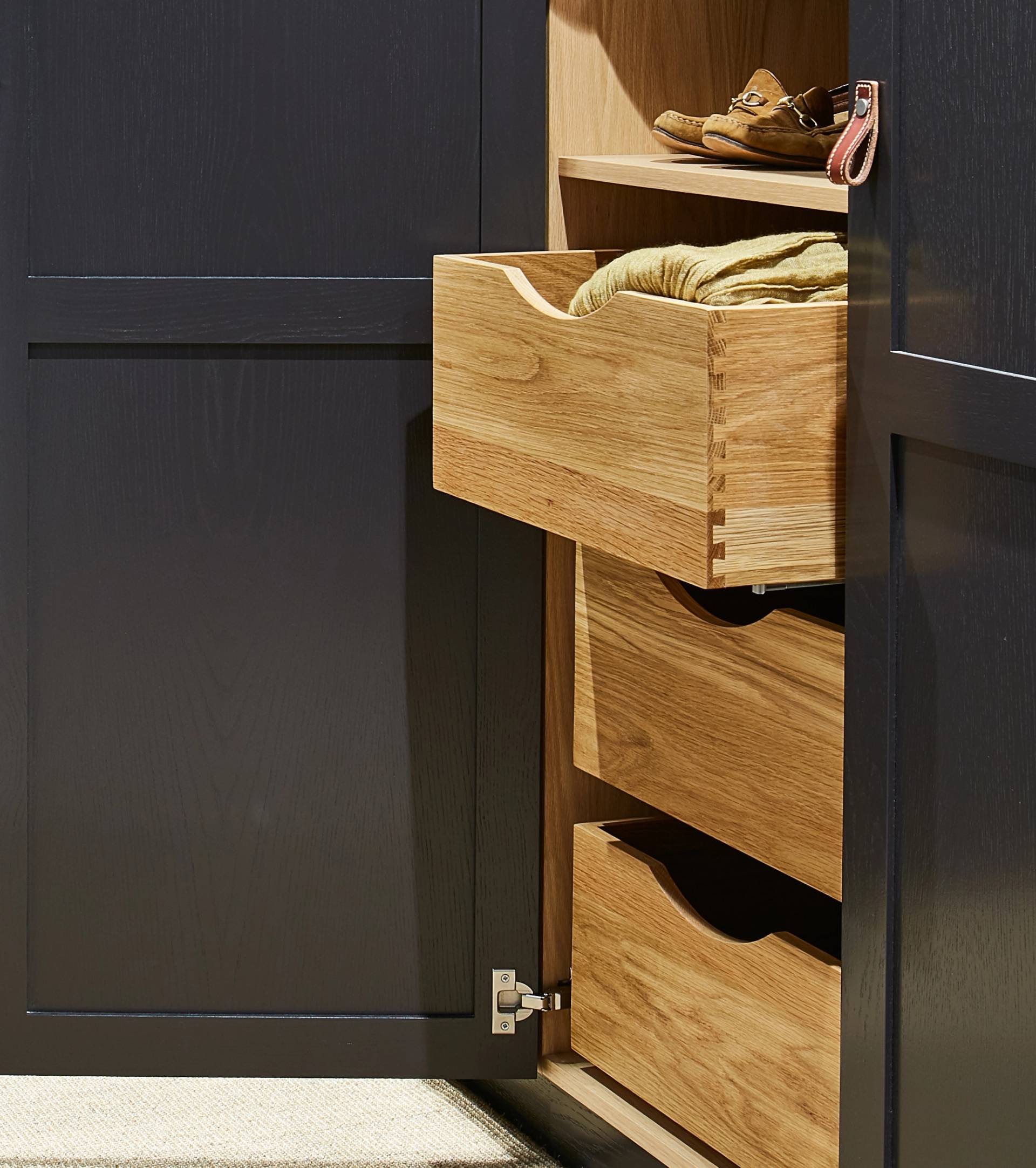 A dark-colored cabinet with its door open, revealing wooden drawers—one partially pulled out with a folded yellow cloth inside. Shoes and a small accessory sit on the top shelf.