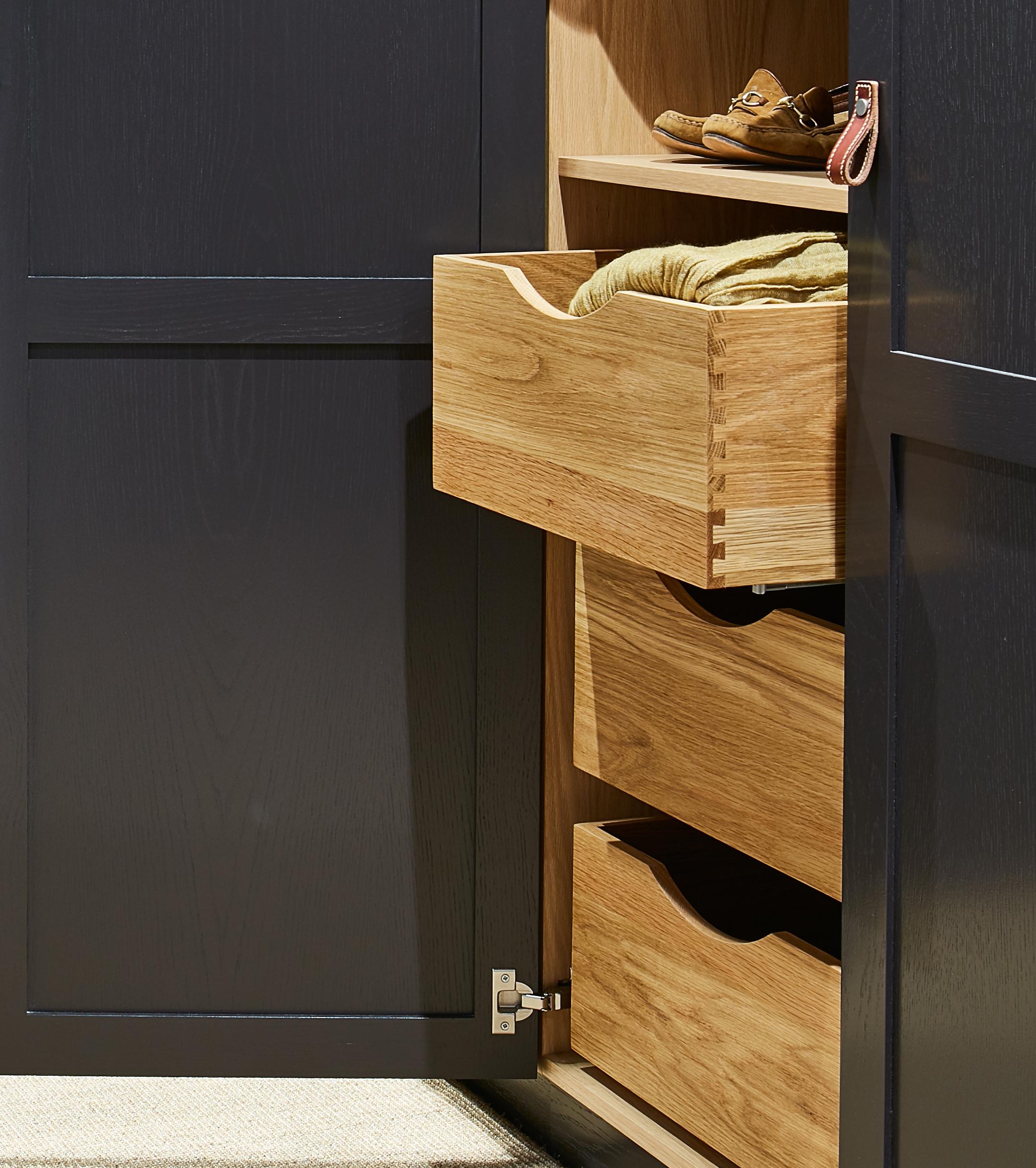 A dark-colored cabinet with its door open, revealing wooden drawers—one partially pulled out with a folded yellow cloth inside. Shoes and a small accessory sit on the top shelf.