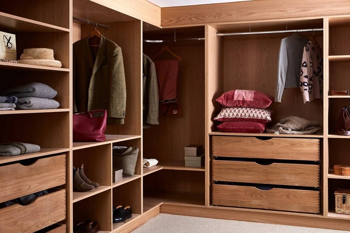 A wooden wardrobe with open shelves and hanging space. It contains folded clothes, a handbag, shoes, and a few hanging jackets and a sweater. There are pillows on one shelf and two drawers underneath with more folded items.