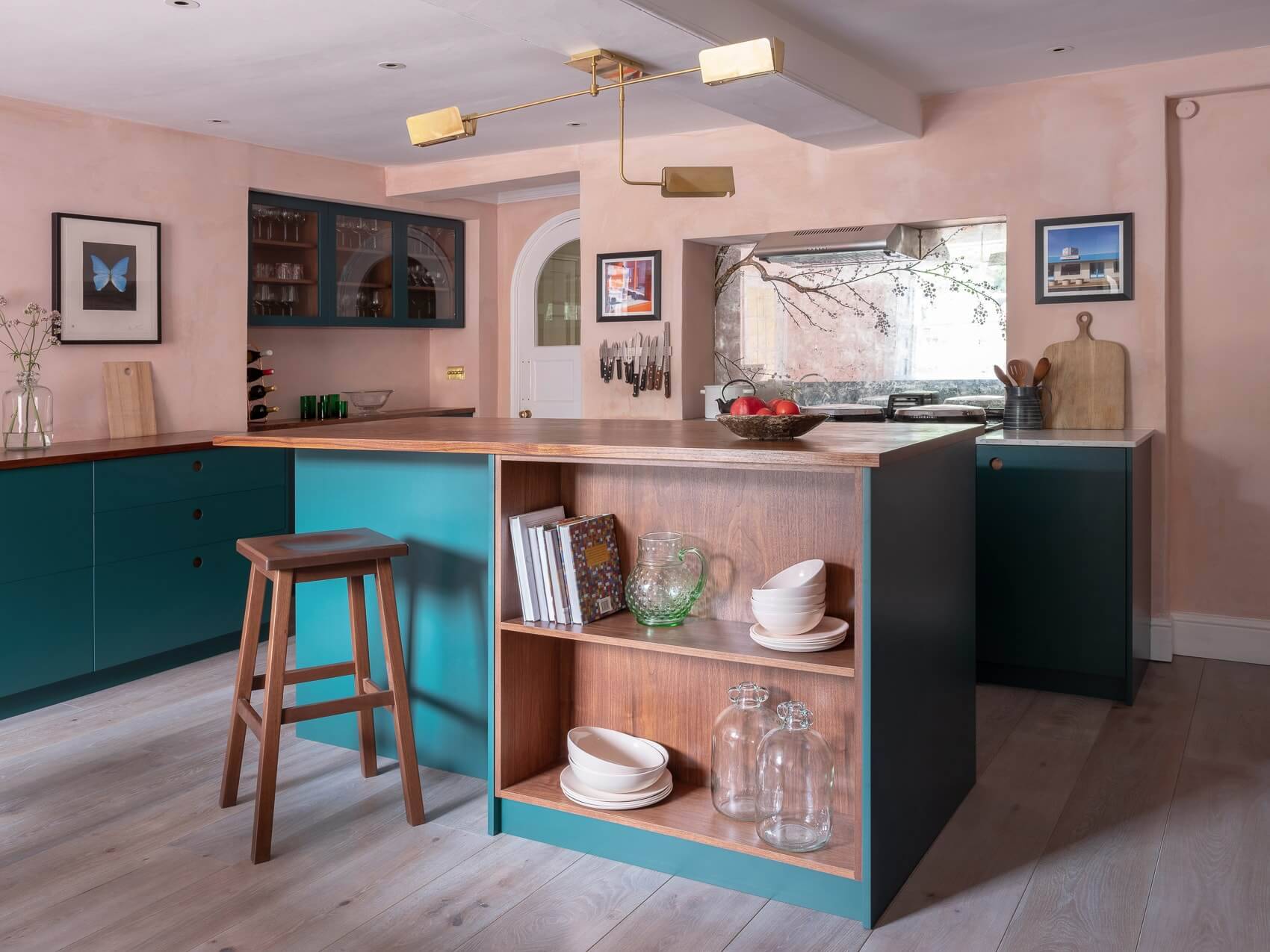 A modern kitchen with teal cabinets, wooden countertops, and open shelving. A stool is by the island, which holds books and dishes. A window lets in light, and there's a geometric ceiling light fixture. Decor includes a framed butterfly art piece.