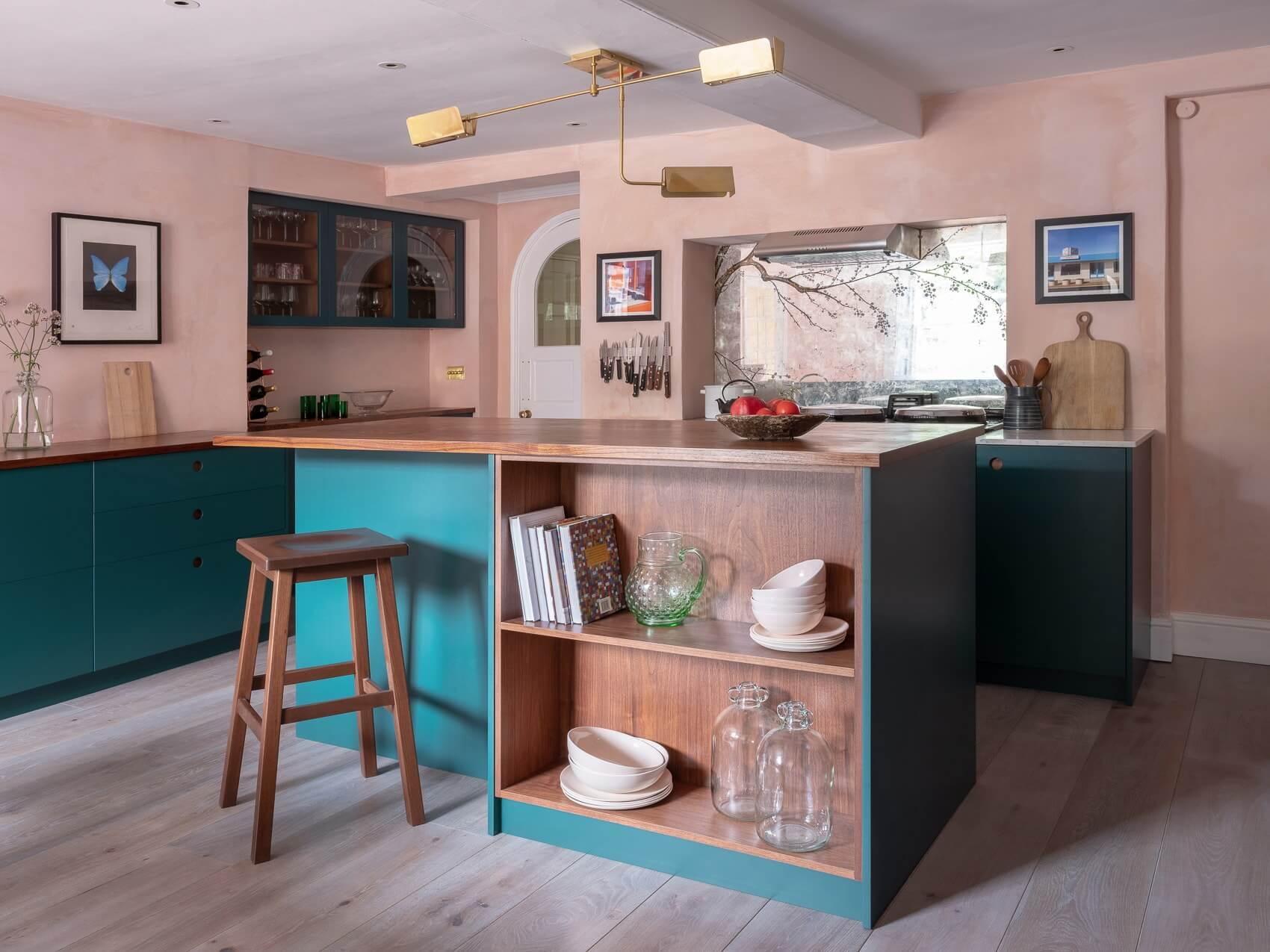 A modern kitchen with teal cabinets, wooden countertops, and open shelving. A stool is by the island, which holds books and dishes. A window lets in light, and there's a geometric ceiling light fixture. Decor includes a framed butterfly art piece.