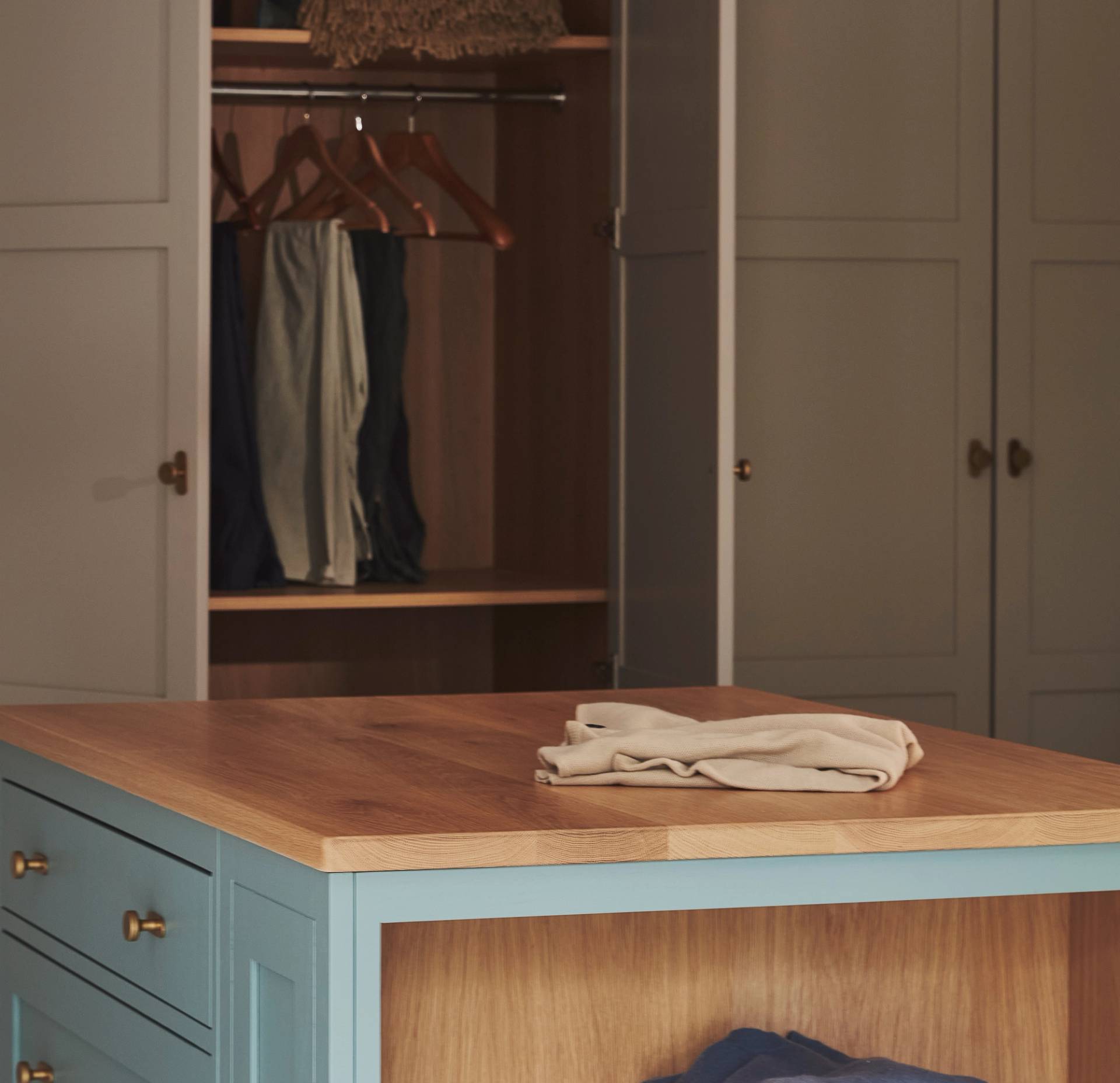 A neatly folded beige shirt sits on a wooden-topped blue dresser in front of an open wardrobe with hanging clothes and empty hangers.