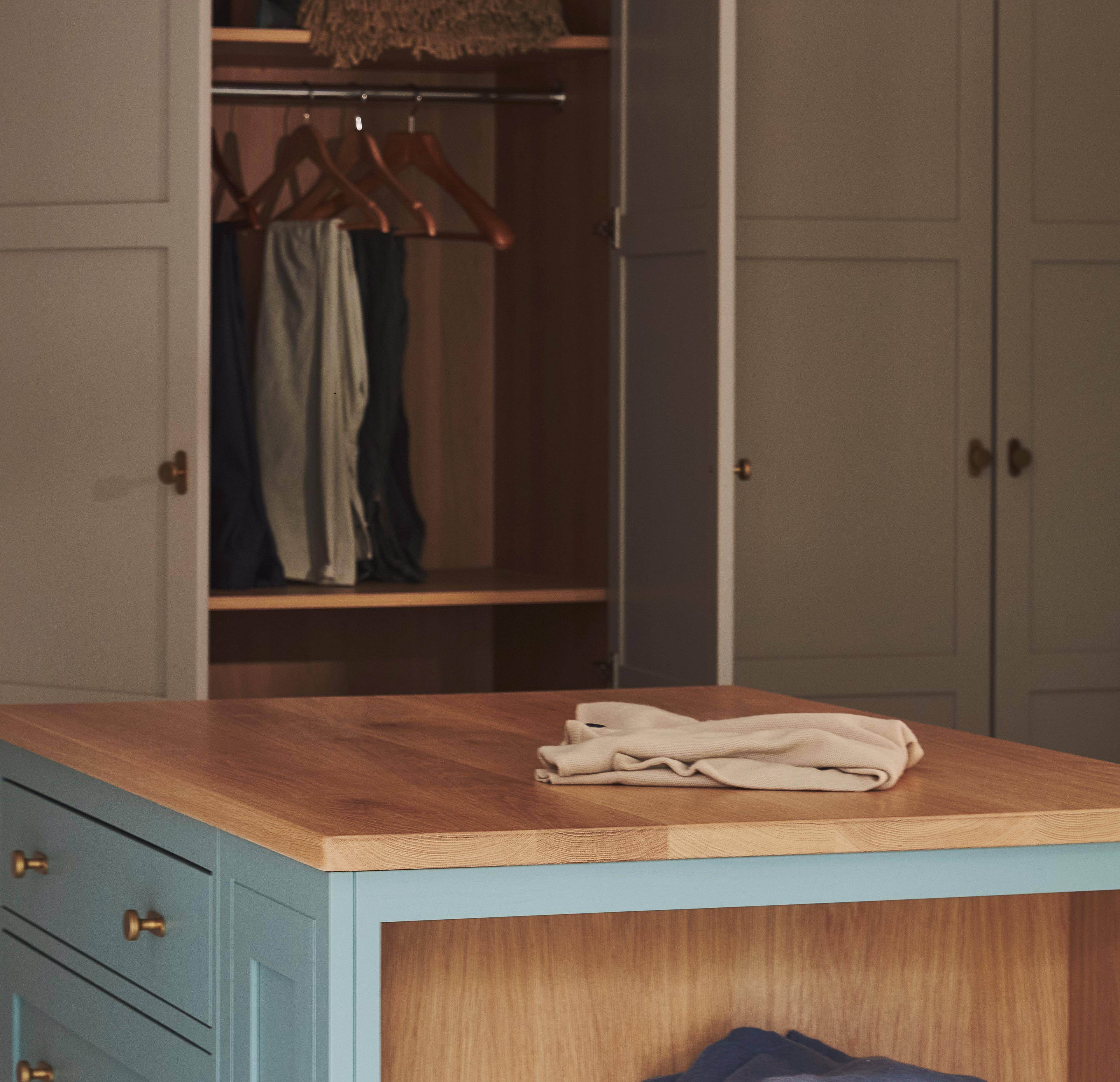 A neatly folded beige shirt sits on a wooden-topped blue dresser in front of an open wardrobe with hanging clothes and empty hangers.