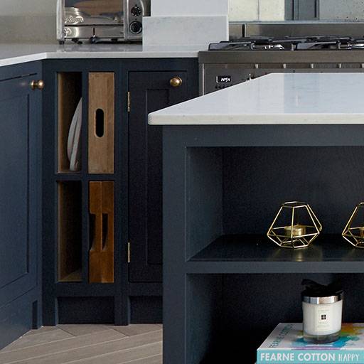 A modern kitchen featuring dark blue cabinets, gold handles, a white countertop, open shelving with books and decor, a gas stove, and a toaster on the counter.
