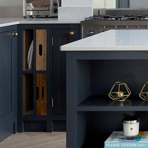 A modern kitchen featuring dark blue cabinets, gold handles, a white countertop, open shelving with books and decor, a gas stove, and a toaster on the counter.