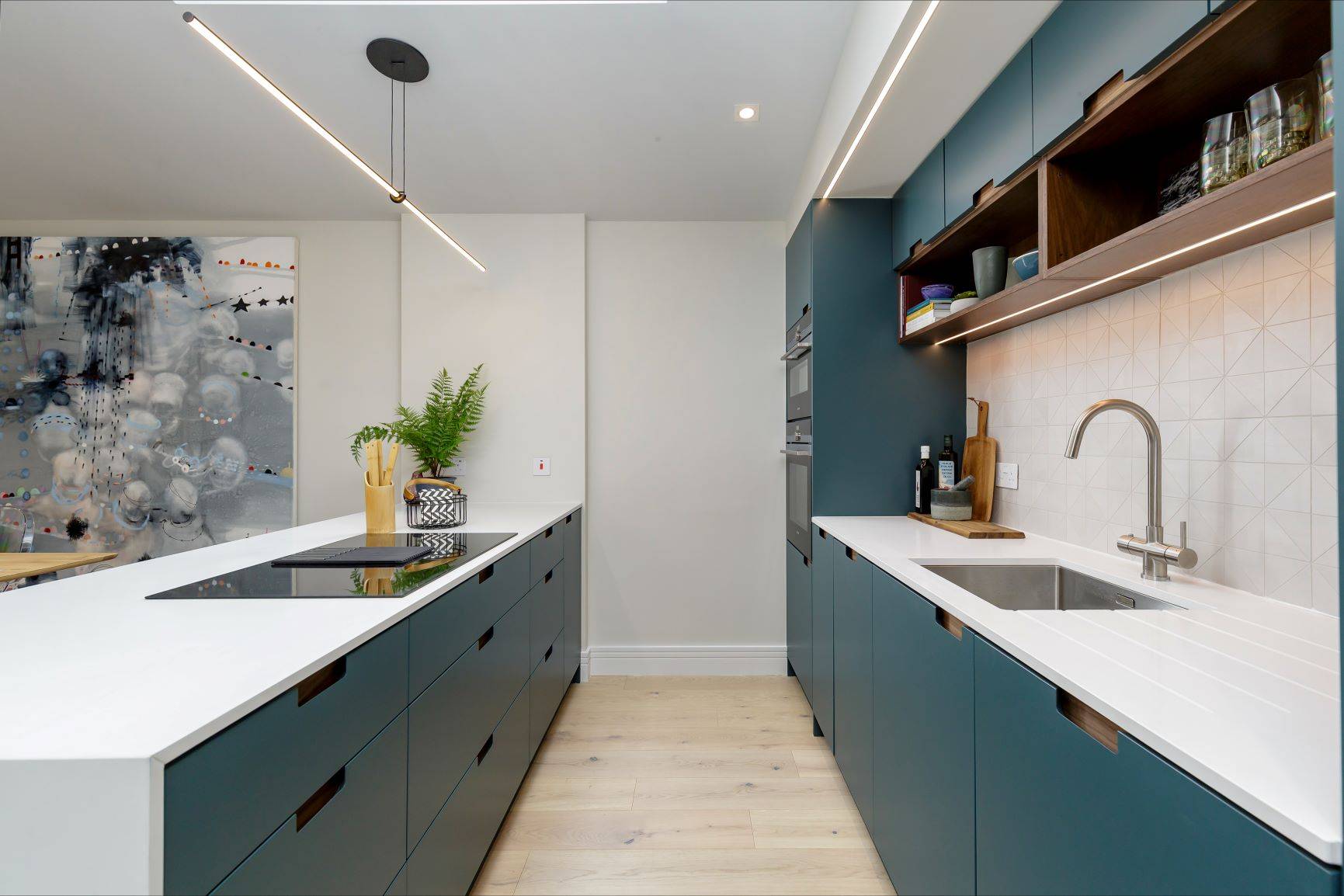 Modern kitchen with sleek blue cabinets, white countertops, and a minimalist design. Features an island with an induction cooktop, an abstract wall art piece, wooden flooring, and under-cabinet lighting. A plant adds a touch of greenery.