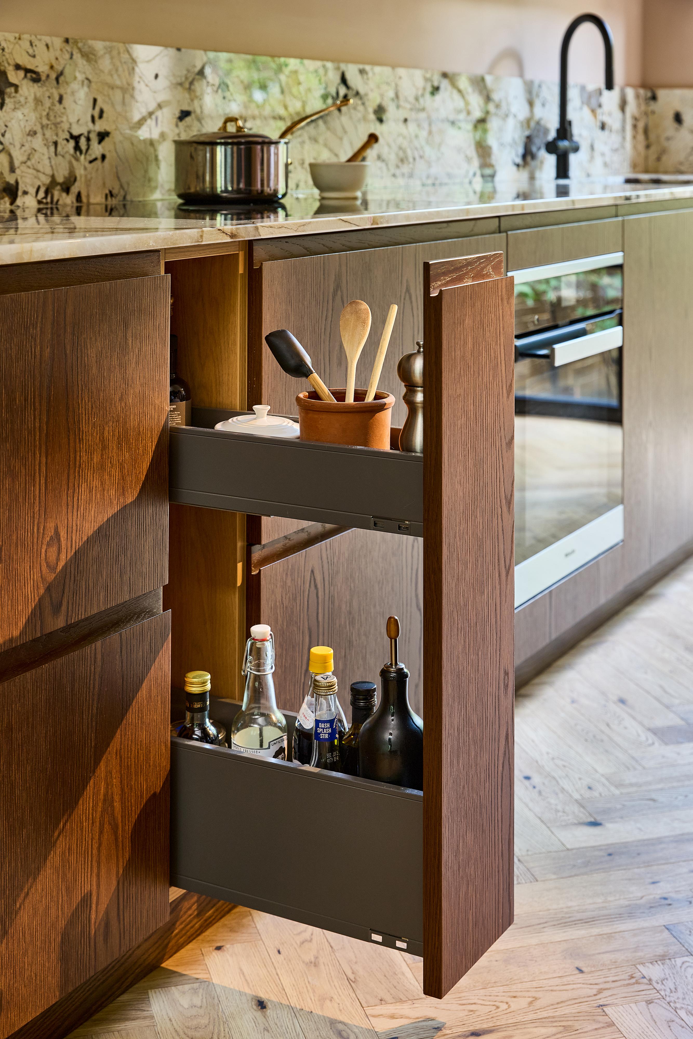 A modern kitchen features a pull-out wooden cabinet with shelves holding utensils, spices, and bottles of oil and vinegar next to an oven and sink.