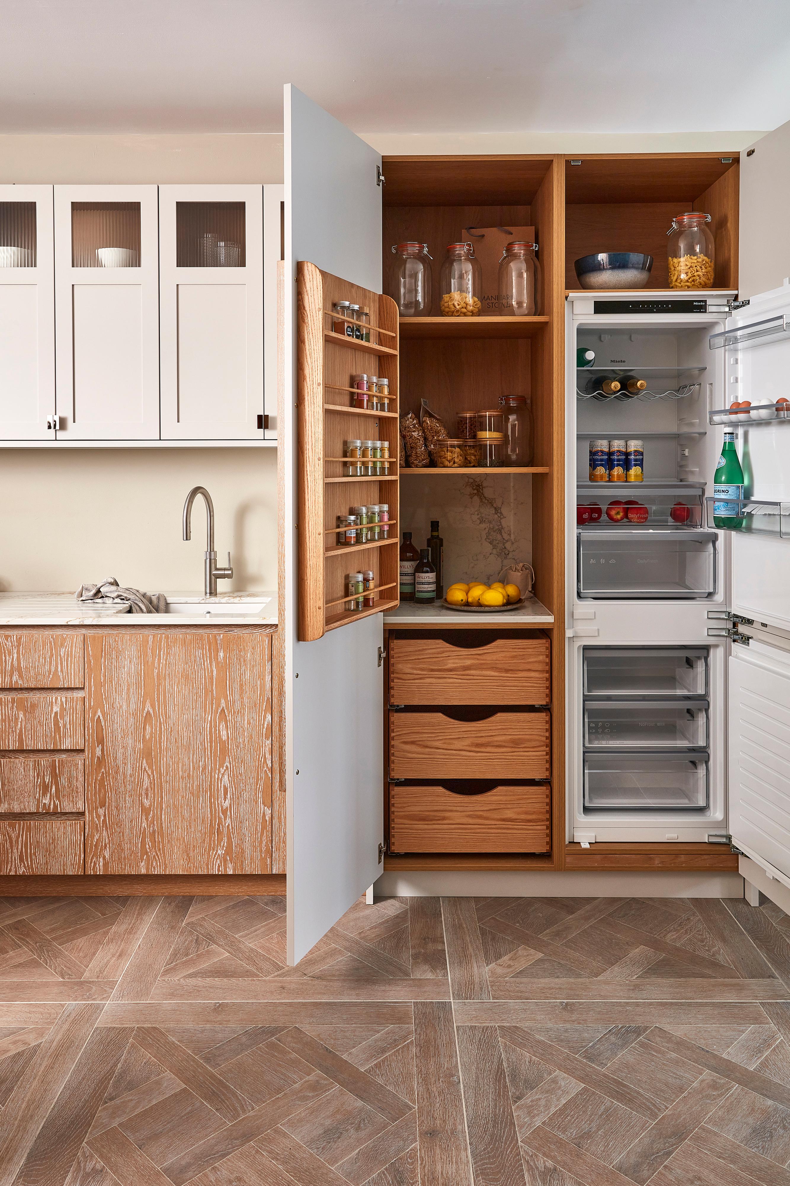 A modern kitchen featuring white upper cabinets with glass inserts, light wood lower cabinets, and a sleek, tall white panelled refrigerator with wooden handles. A stainless steel sink with a faucet is set into the countertop. The floor is parquet wood.